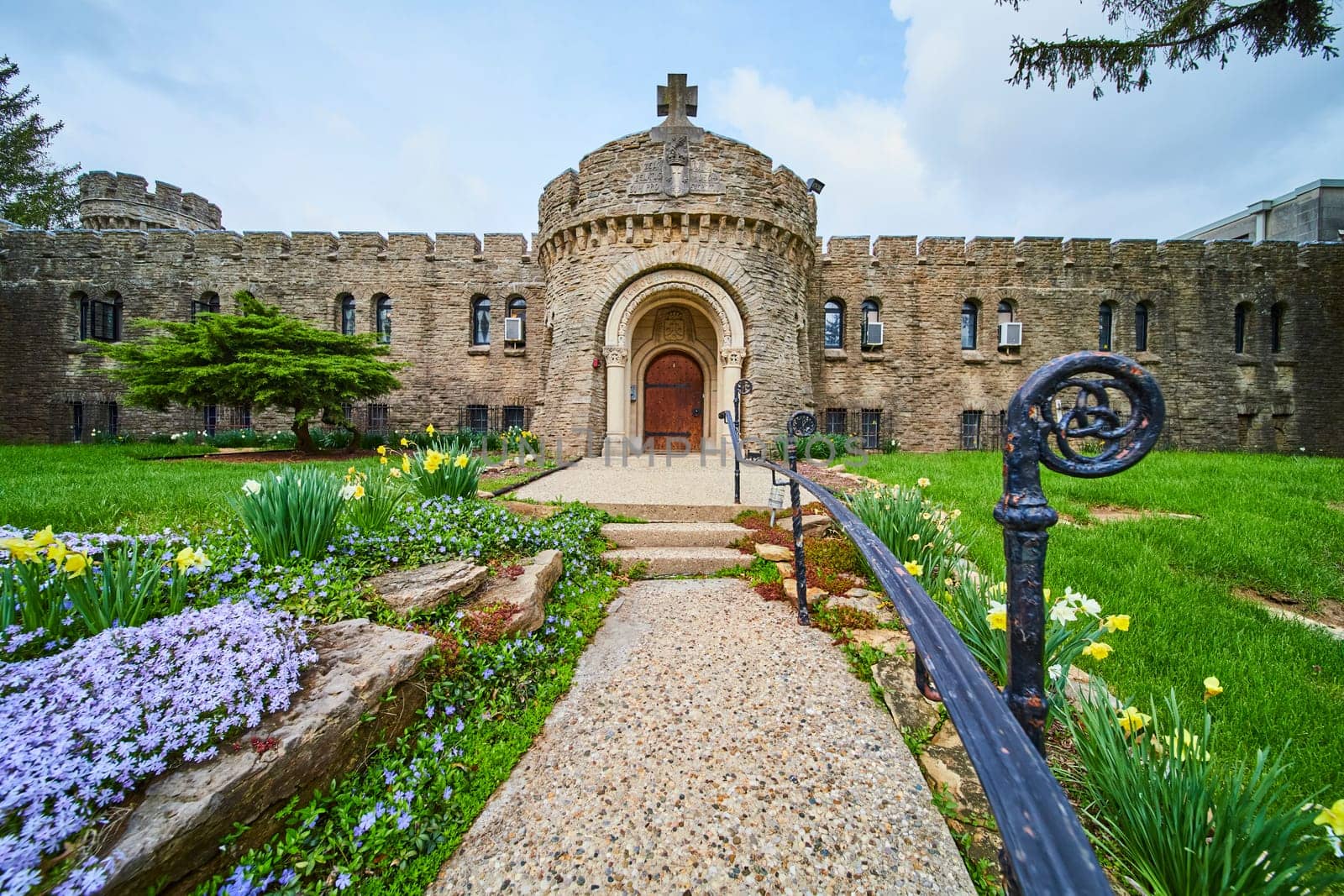 Bishop Simon Brute College, Indiana - A vibrant spring view of a historic castle-like seminary with medieval architecture, along a floral garden path.