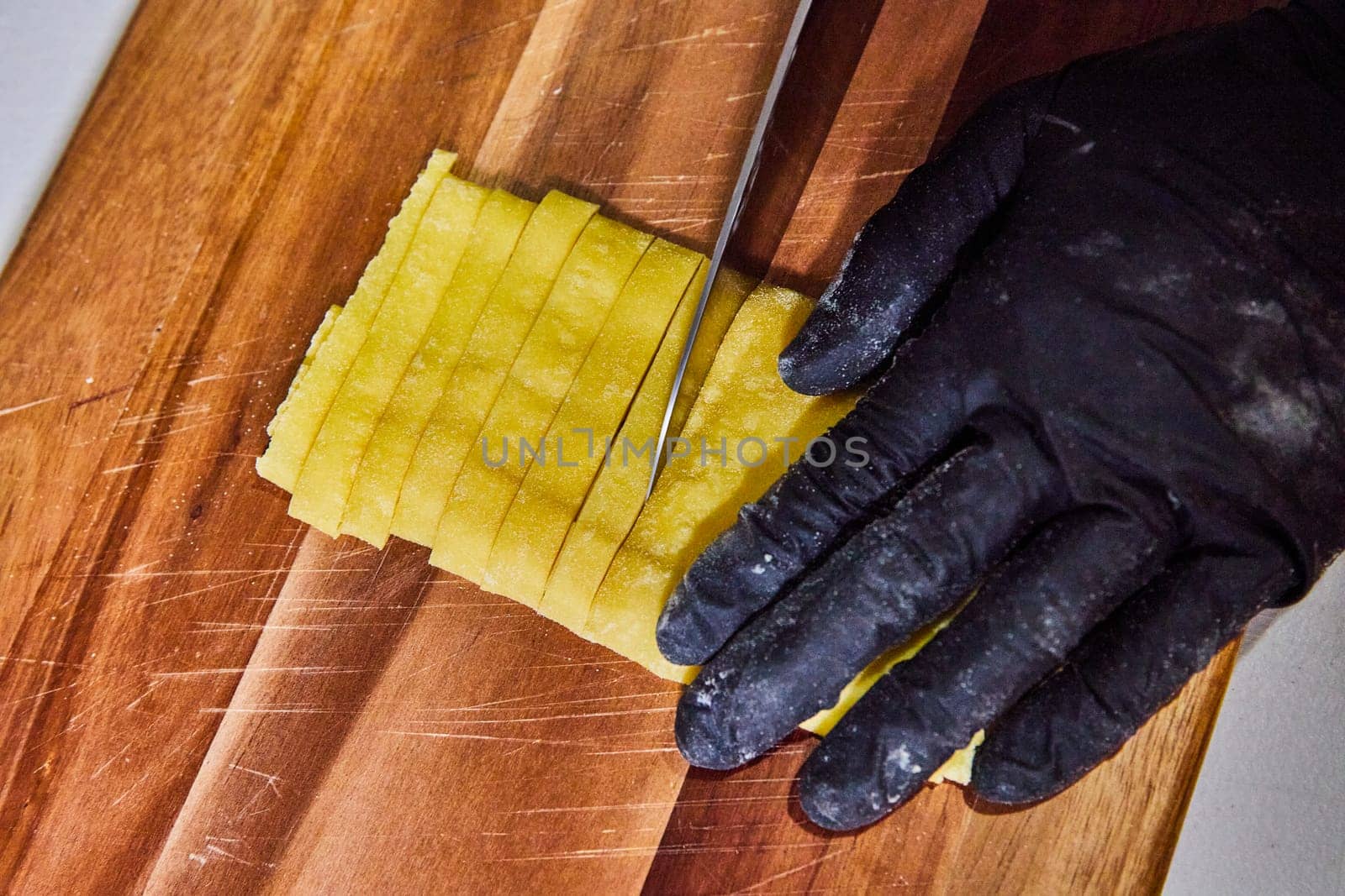 Close-up of gloved hand expertly slicing fresh, yellow homemade fettuccine on well-used wooden board in Indiana kitchen.