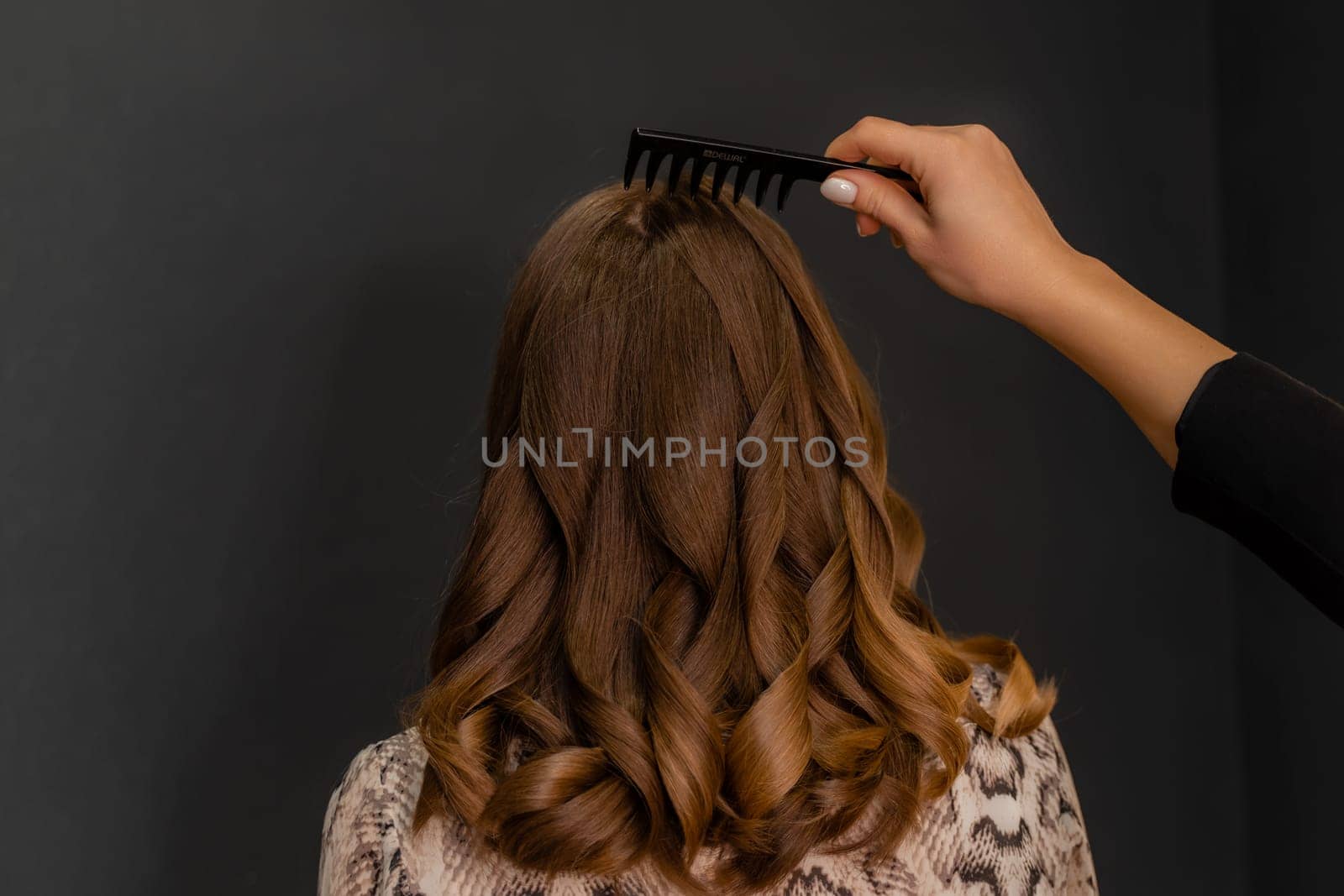 A woman with long brown hair is getting her hair brushed by another person. The brush is black and the woman's hair is curly