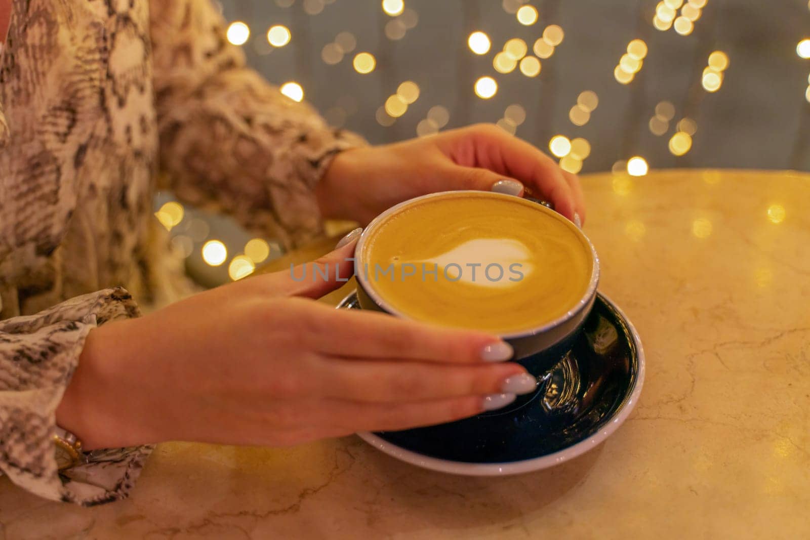 Close up cup of latte coffee in a coffee shop. Female hands hold a cup of coffee with foam latte in the shape of a heart on a table in a cafe. On the background, the lights are in blur