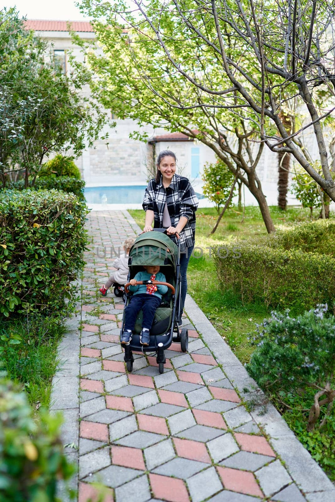 Smiling mother pushing little girl in stroller along path in garden. High quality photo