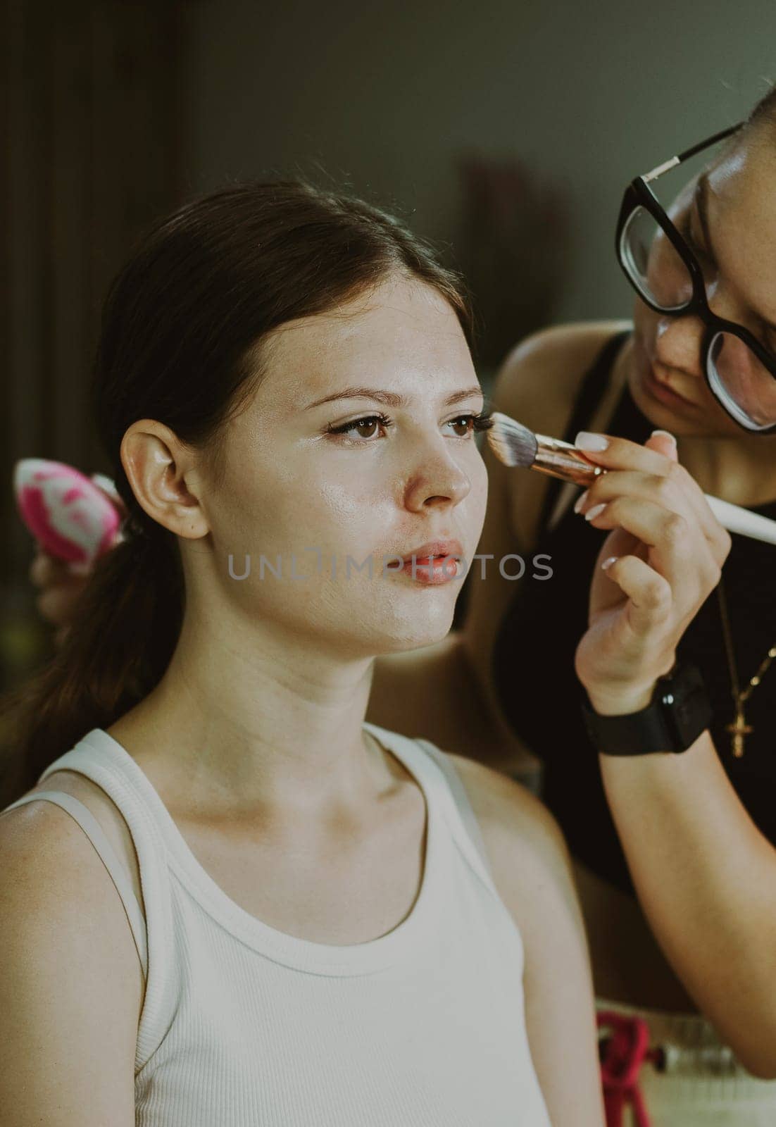 One young handsome Caucasian makeup artist applies blush with a brush to the cheekbones of a girl sitting in a chair early in the morning in a beauty salon, close-up side view. Step by step.