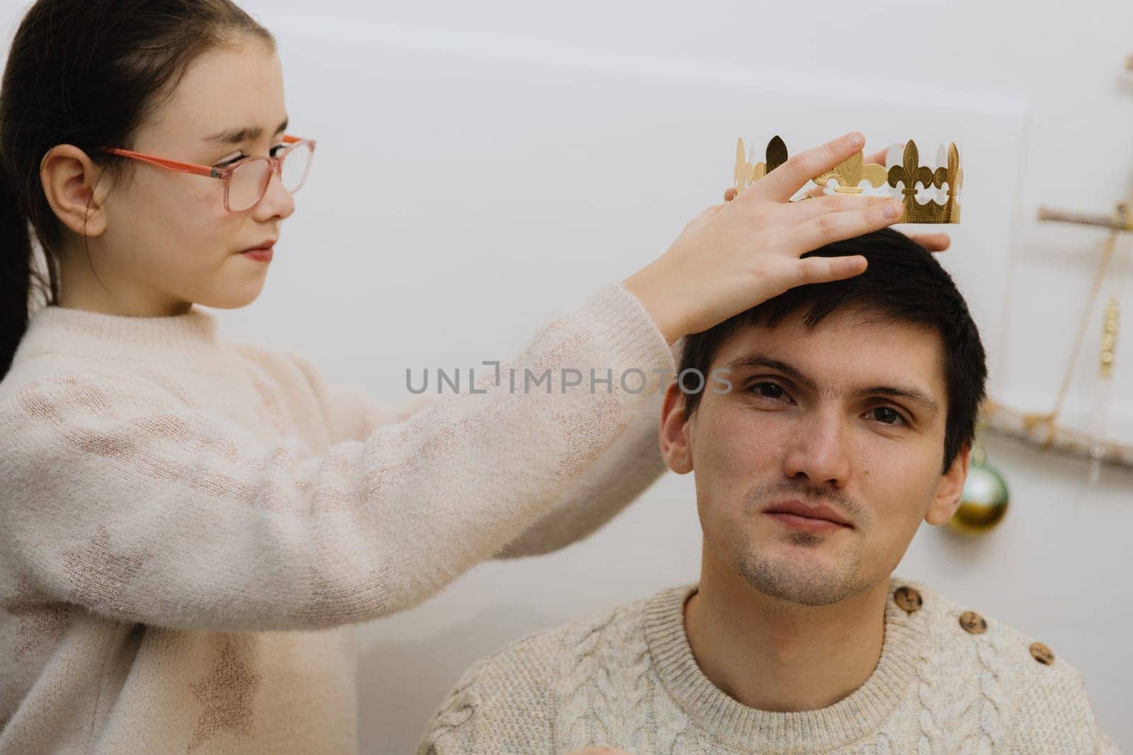 One beautiful Caucasian girl with glasses solemnly puts a golden paper crown on an adult guy who ate a royal galette and found a gift while sitting at the table in the kitchen, close-up side view.