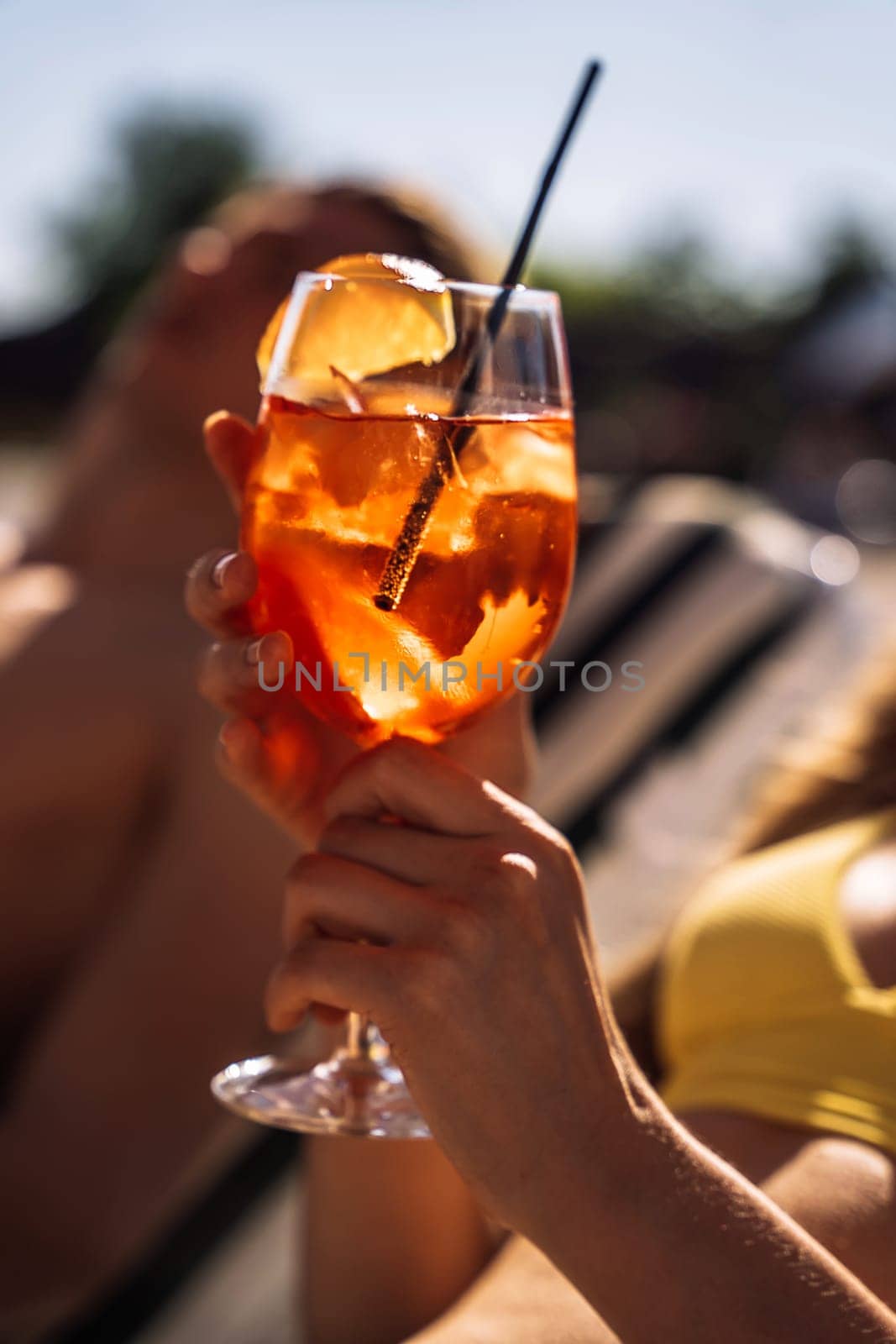 Closeup of young womans hand with neat red manicure holds elegant glass with long stem of aperol. Tasty citrus cocktail with orange slice. Sunny day, spa resort.