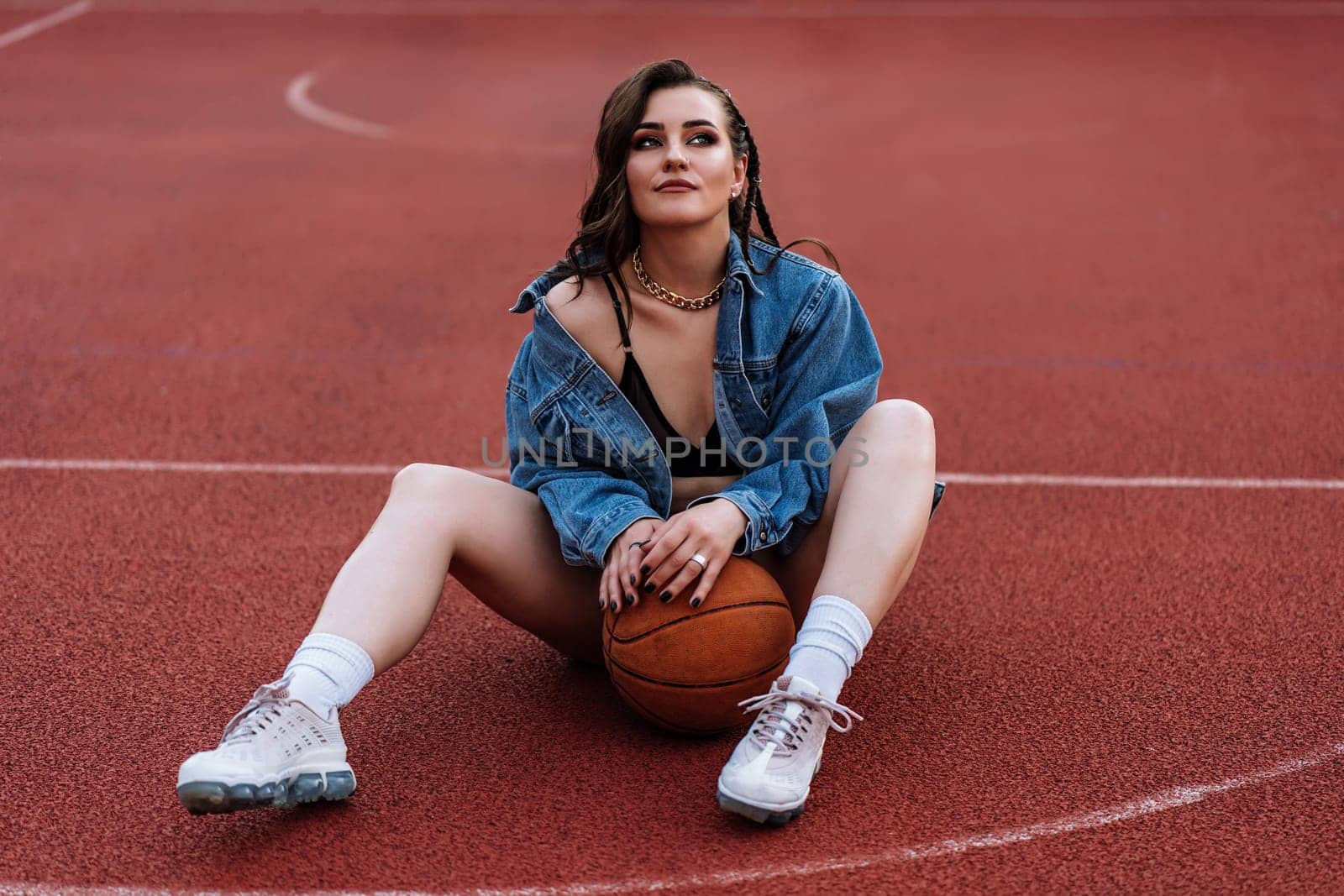 Outdoor portrait of dreaming girl in bra and jacket posing on the ground. SWAG concept. Excited brunette woman sitting at basketball court with ball between her legs. by OnPhotoUa