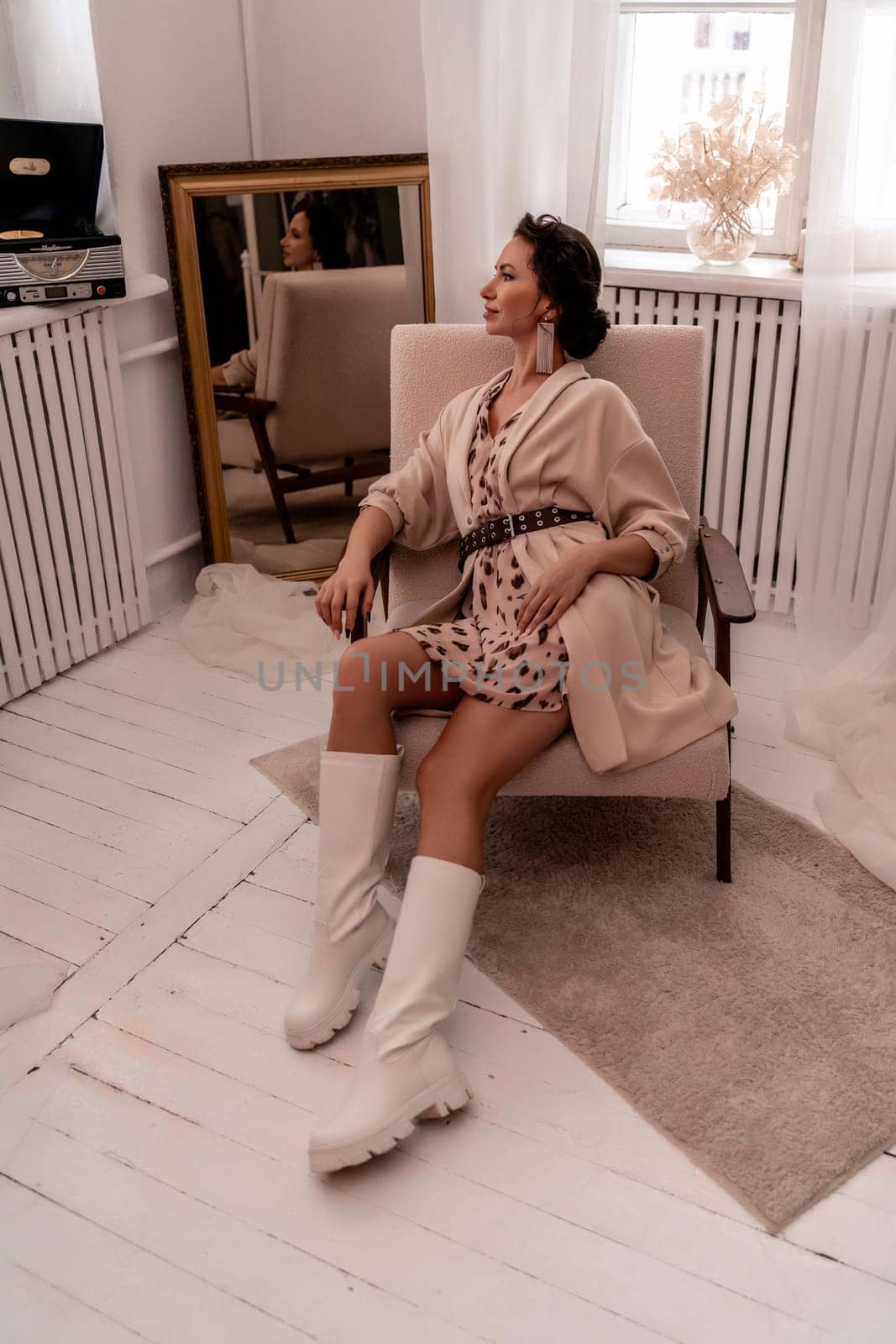 A woman is sitting in a chair wearing a white dress and white boots. She is wearing a brown jacket and a brown belt