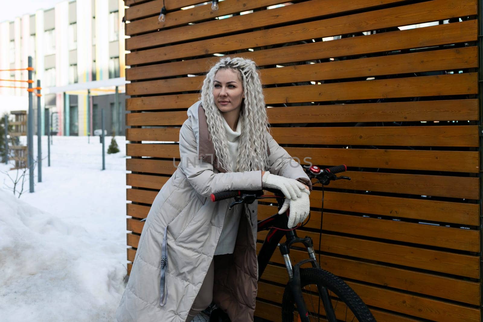 A woman is standing next to a bicycle on a snow-covered ground. She is wearing winter clothing and looking ahead. The bike is parked in a snowy landscape.