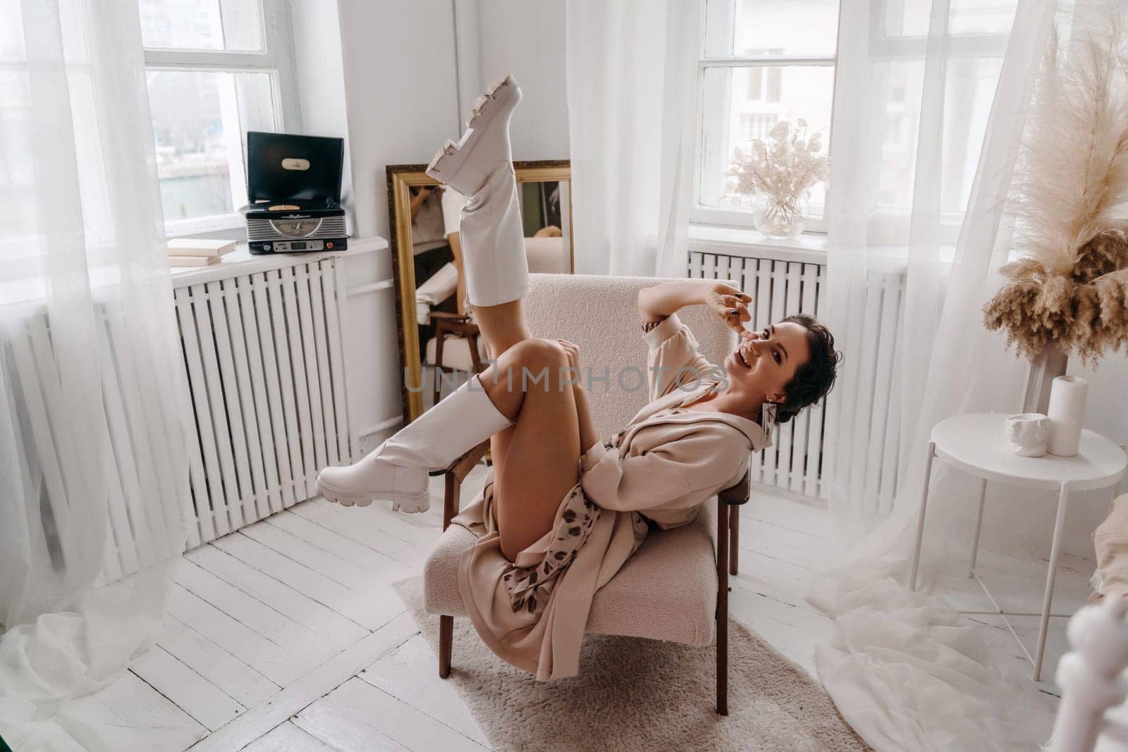A woman is sitting in a chair with her legs spread apart. She is wearing white boots and a white dress. The room is decorated with a white color theme