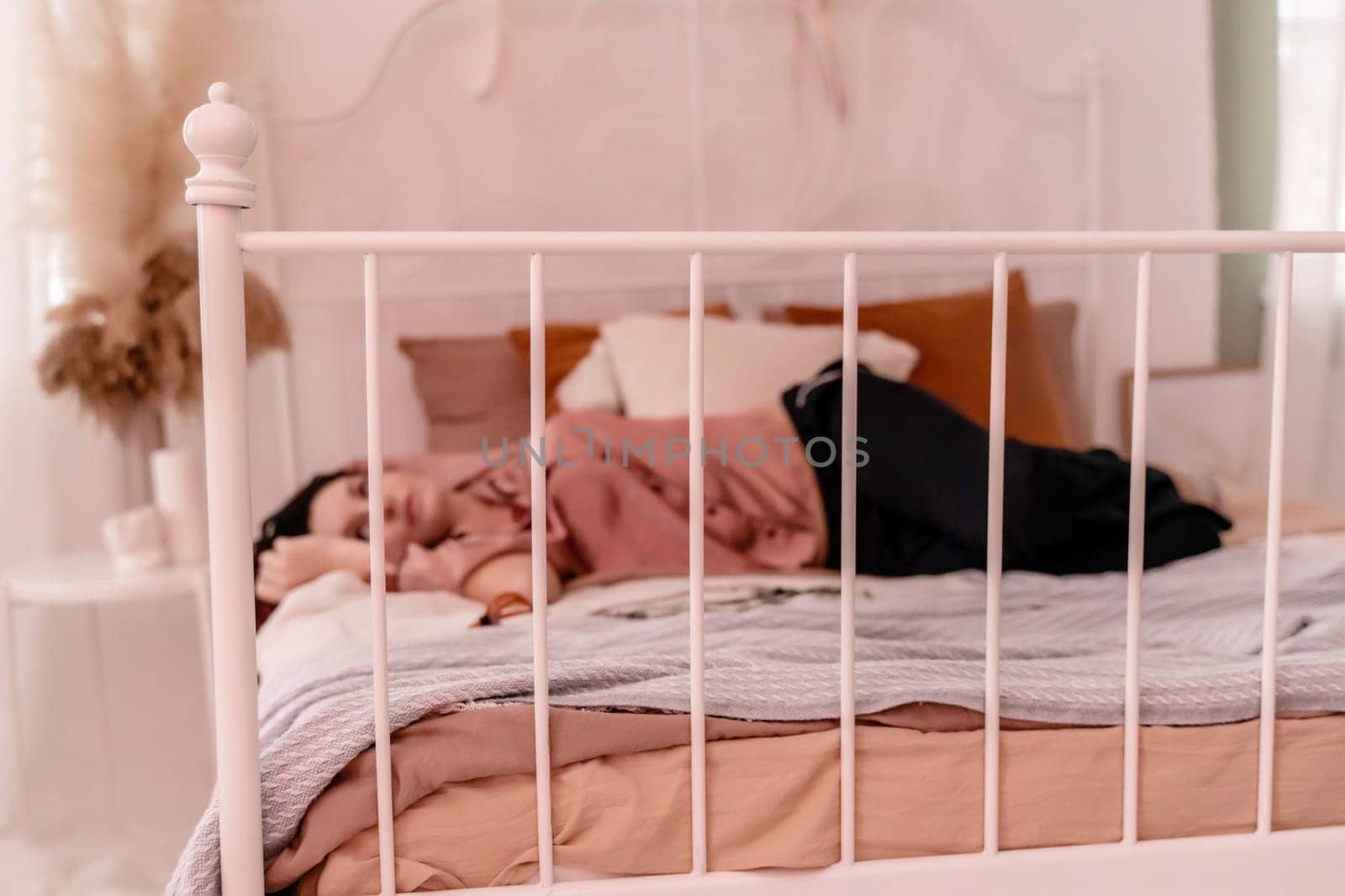 A woman is laying on a bed with a white metal frame. She is wearing a pink shirt and is resting her head on her hand. The bed is covered in a white blanket and pillows
