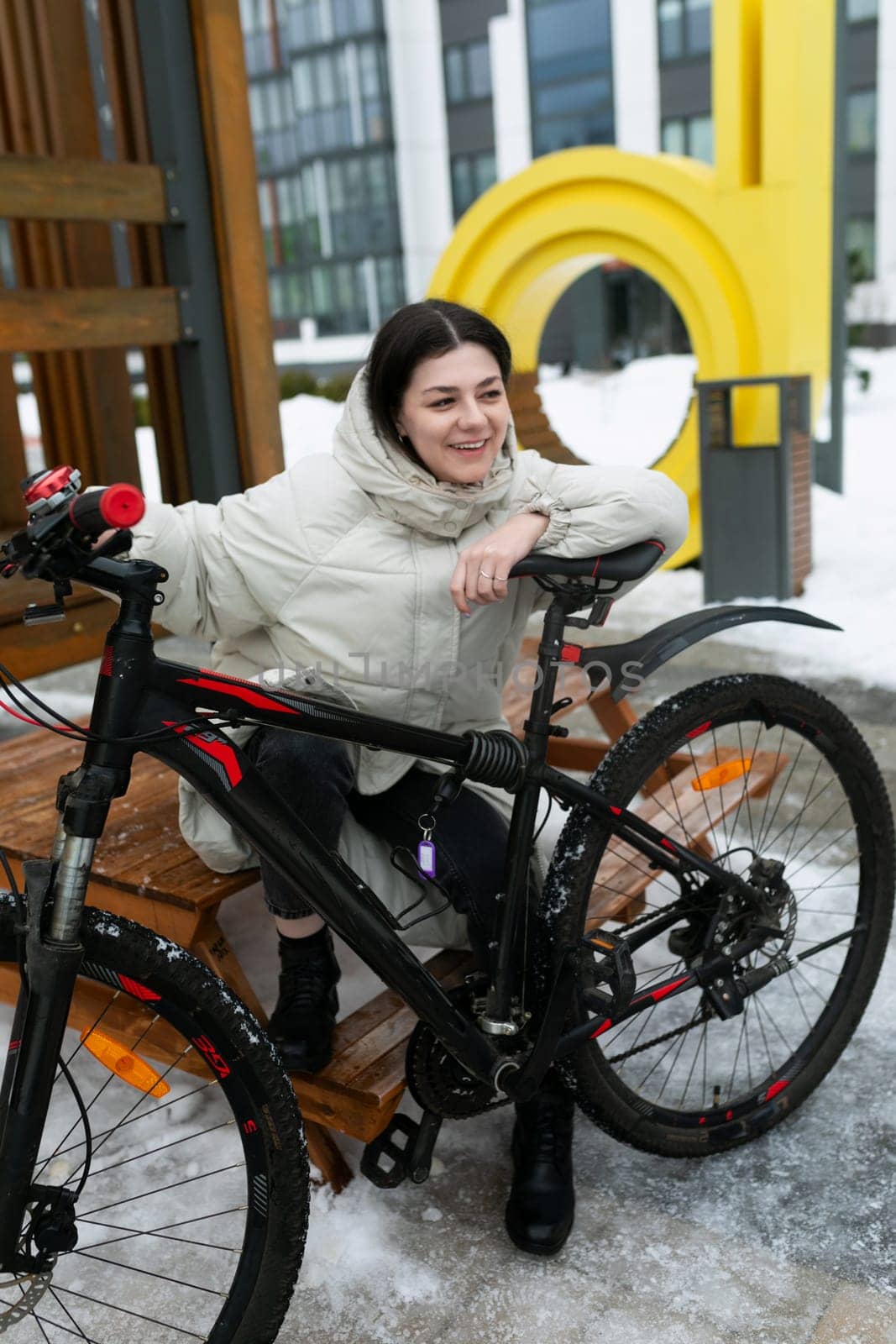 A woman is seated on a bench outdoors, next to a bicycle. She appears relaxed, gazing into the distance. The bike is propped up beside her, adding to the scene of urban leisure.