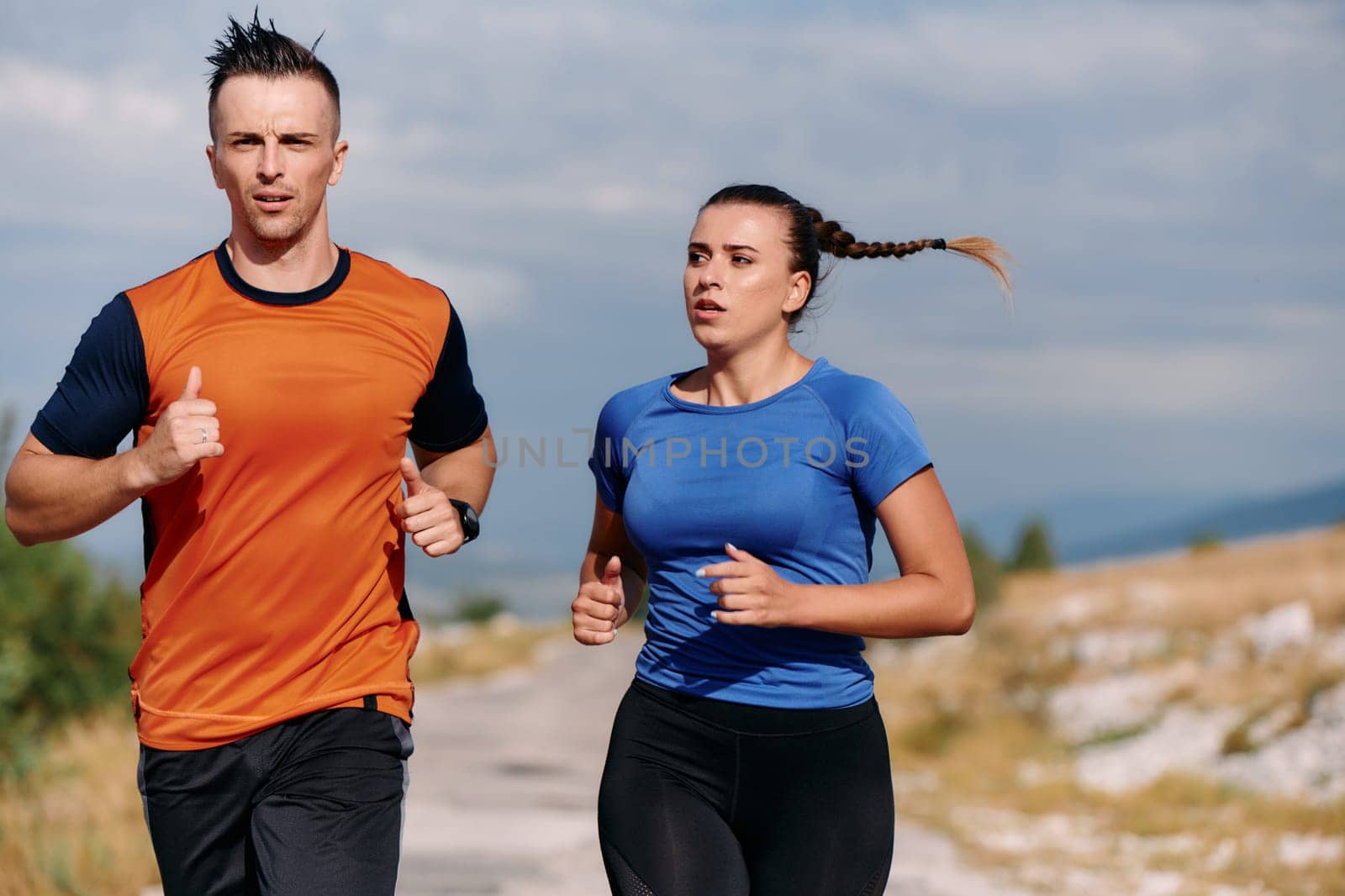 A couple dressed in sportswear runs along a scenic road during an early morning workout, enjoying the fresh air and maintaining a healthy lifestyle.