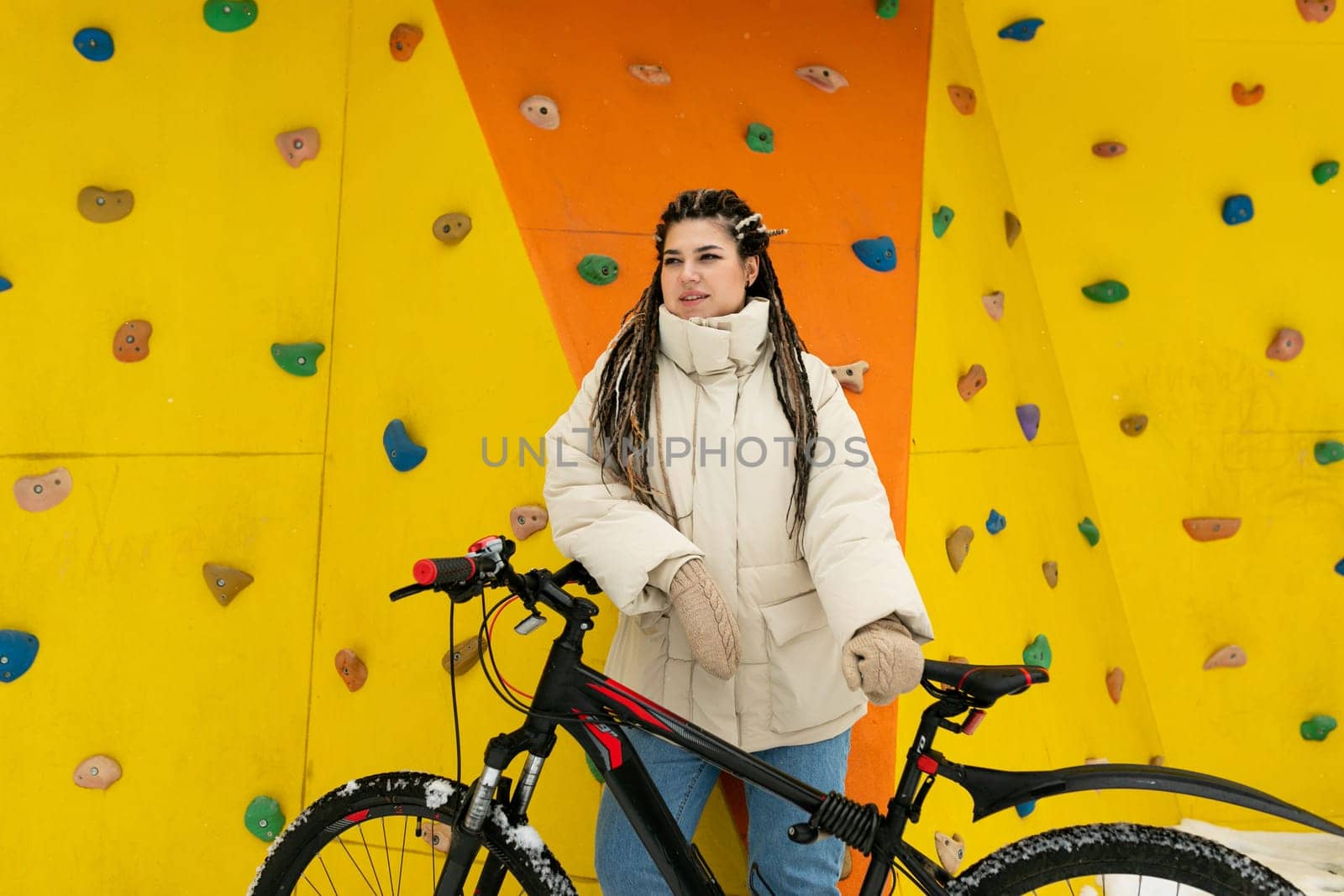A woman stands next to a bicycle in front of a towering climbing wall. She appears ready for a physical challenge, with climbing gear visible. The scene suggests a combination of outdoor activity and adventure.