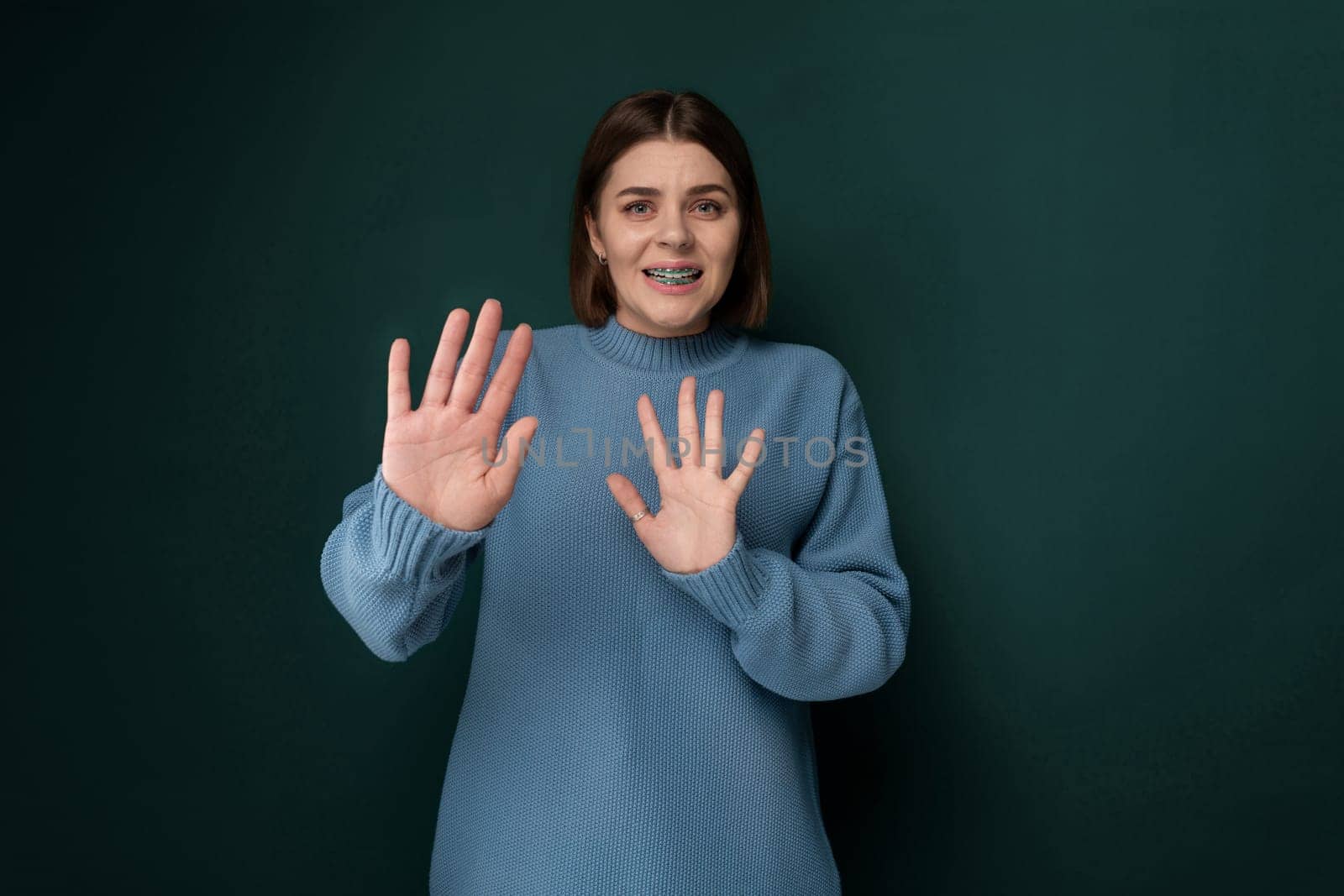 A woman wearing a blue sweater is standing with her hands raised up. She appears to be in a pose of surrender or excitement, with a neutral expression on her face.