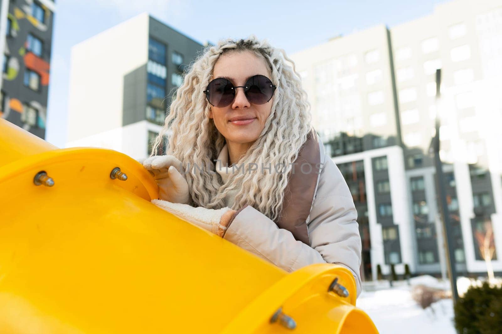 A woman wearing sunglasses is leaning casually on a vibrant yellow structure. She appears relaxed and contemplative as she stands against the structures bright color.