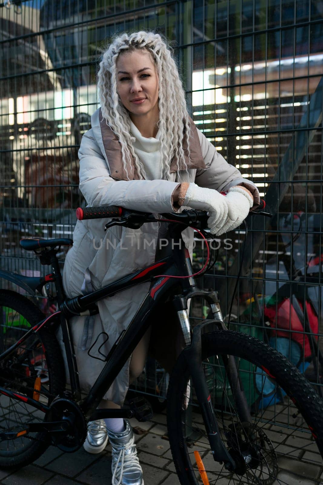 A woman is casually sitting on top of a bike parked next to a fence. She appears relaxed and is looking around. The fence is made of wood and separates a grass field from the sidewalk.
