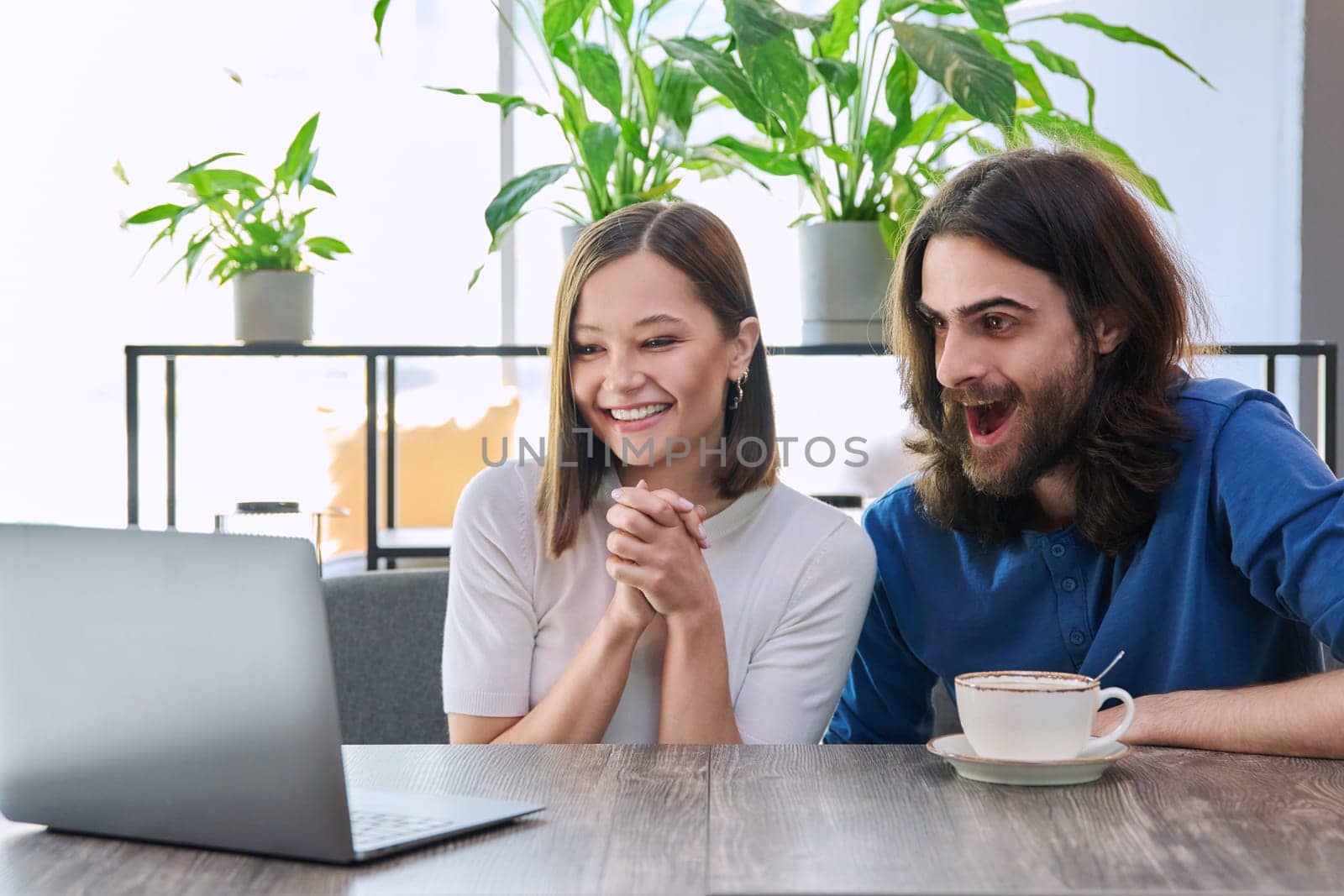 Happy surprised young couple looking at laptop together while sitting in cafeteria by VH-studio