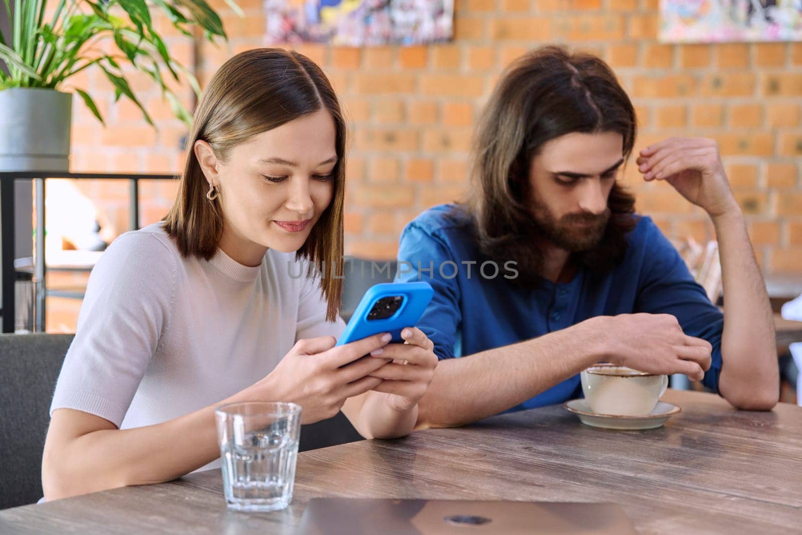Young people, couple man and woman relaxing together in cafe, using smartphone by VH-studio