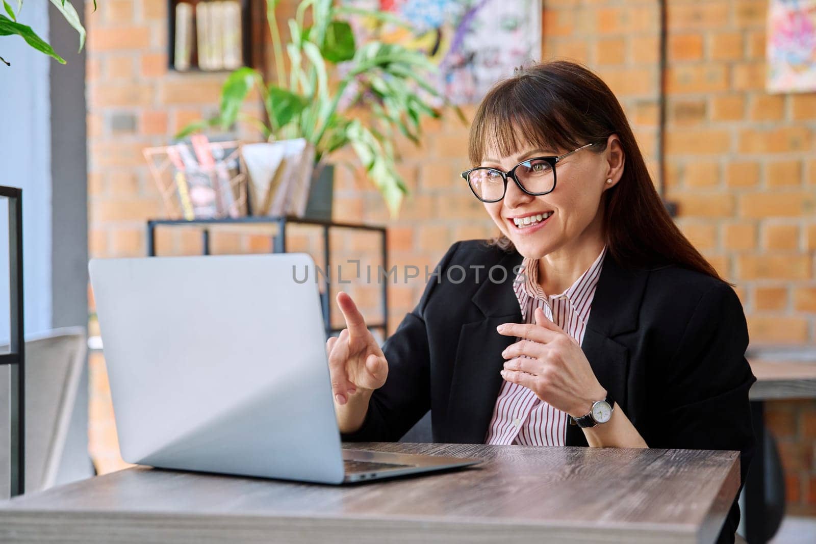 Cheerful talking middle-aged woman having video call using laptop in coworking cafe by VH-studio
