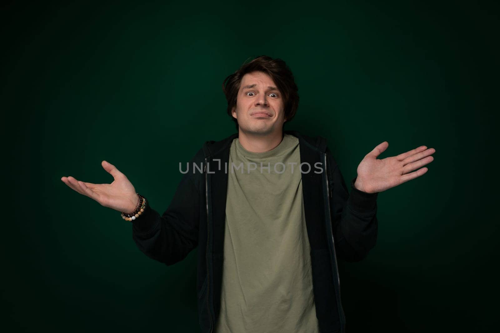 A man stands in front of a solid green wall, looking straight ahead with a neutral expression. He is wearing casual clothing and appears to be waiting or posing for a photograph.