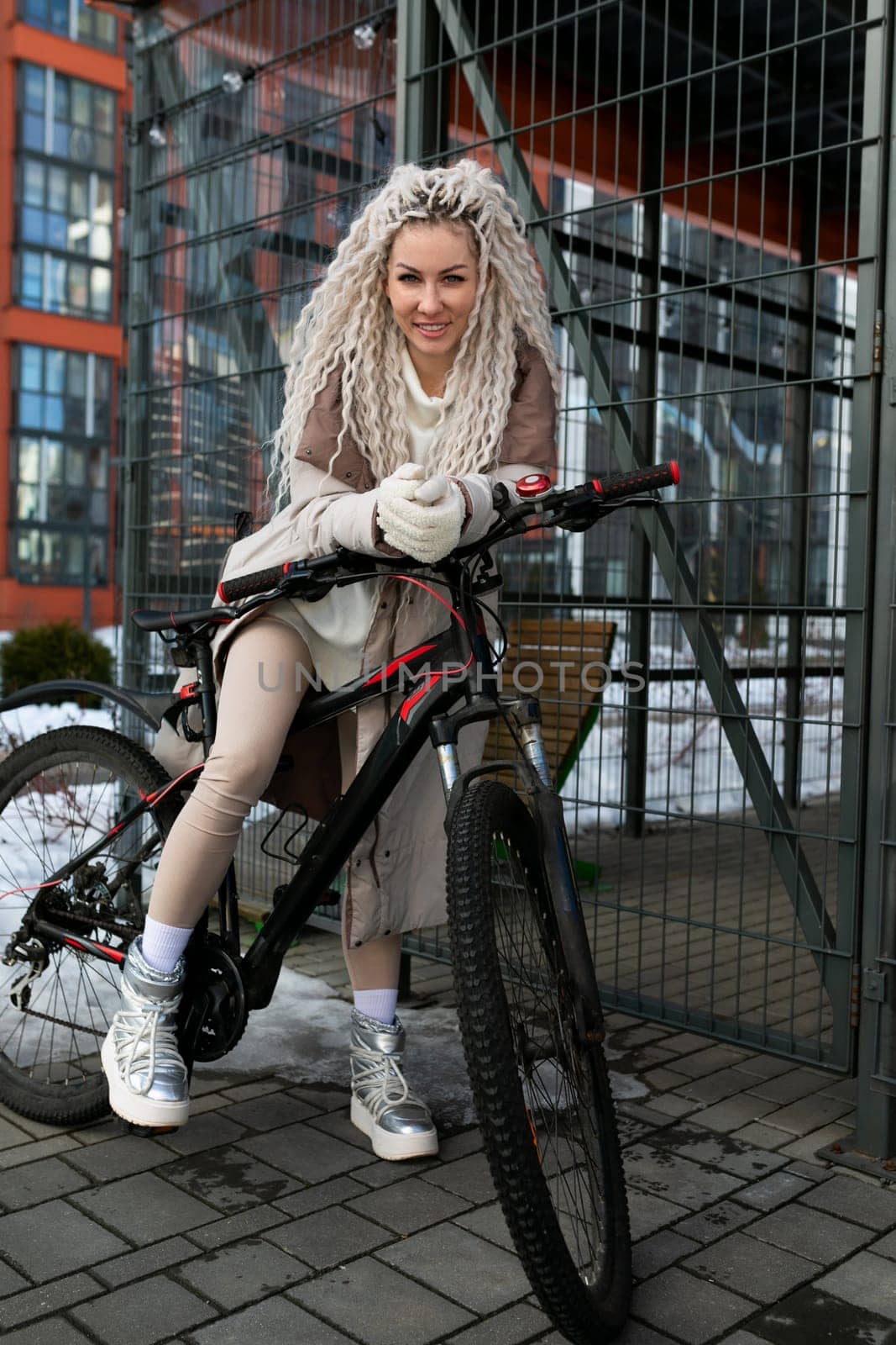 A woman sitting atop a stationary bike positioned next to a metal fence. She appears relaxed and casual, with her feet firmly planted on the ground. The bike is parked on a sidewalk, and the fence extends vertically in the background.