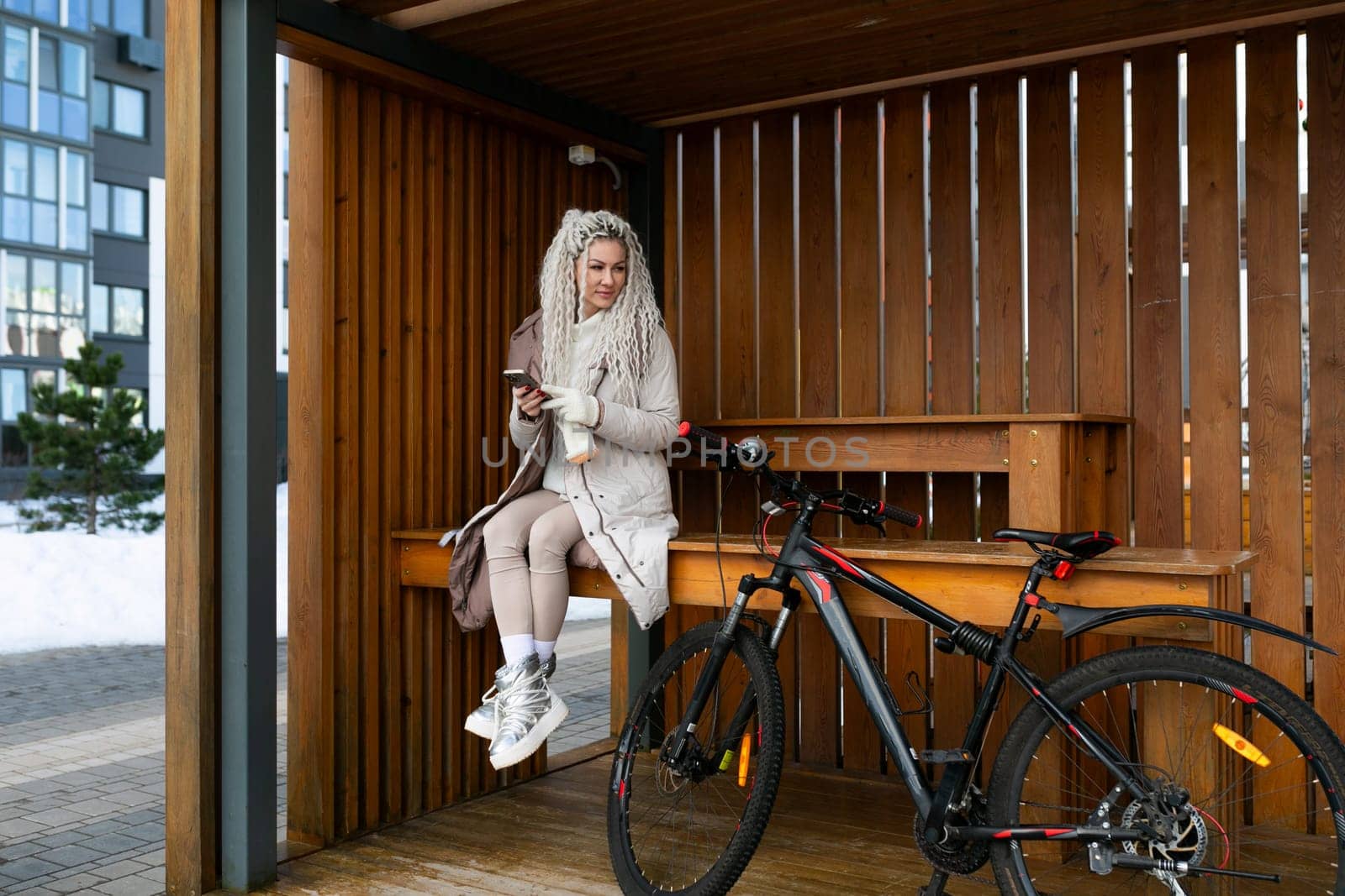 A woman in casual attire sitting on a wooden bench beside a silver bicycle. The woman is looking ahead with a relaxed posture, surrounded by a park setting with greenery in the background.