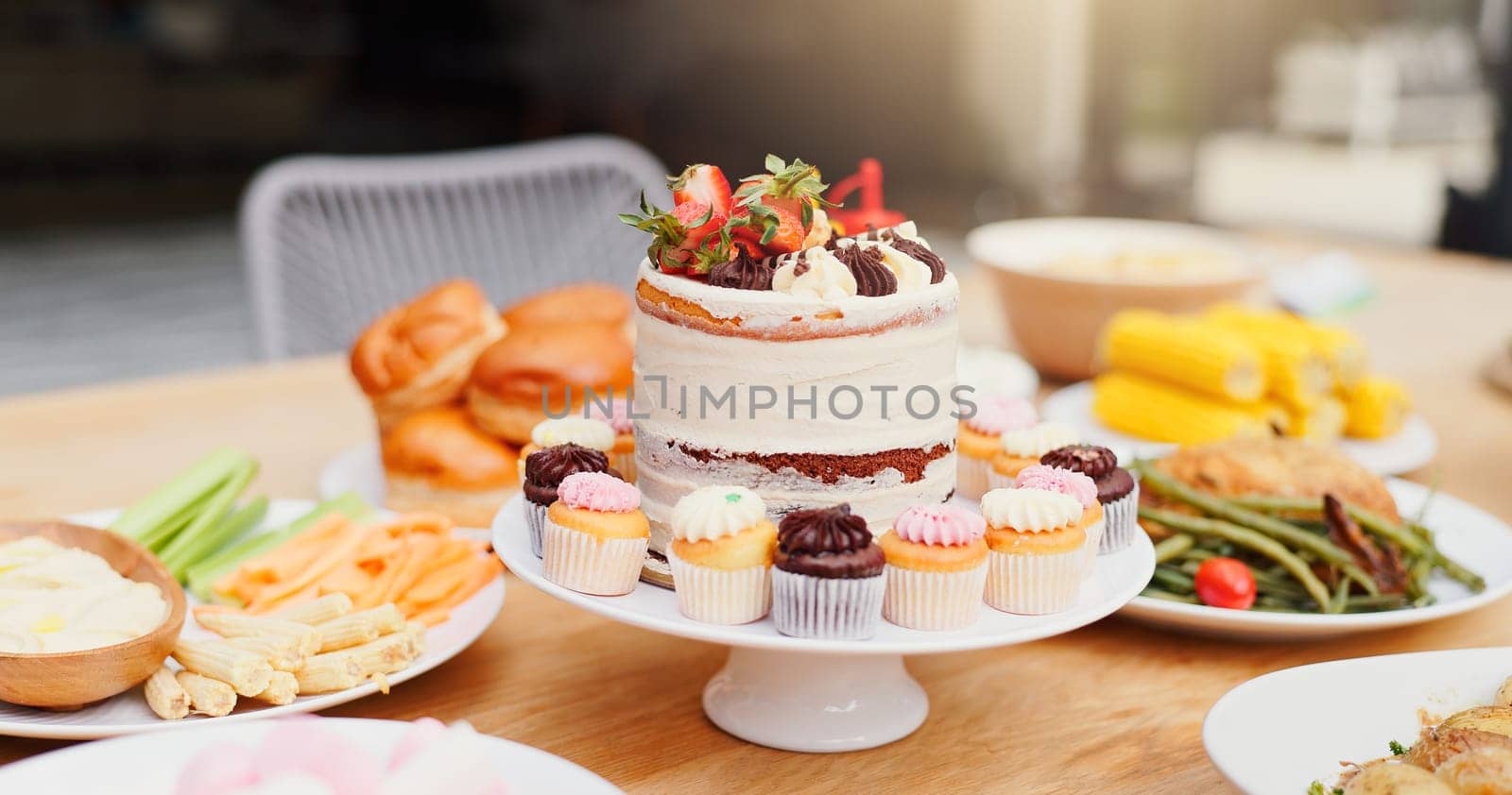 Birthday cake, table and food for event, celebration and party on patio with lunch meal. Dessert, home and outdoor with feast, vegetable and sweet baking with cupcake and sugar snack on a plate.