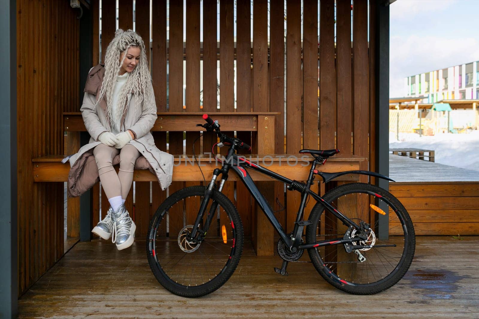 A woman is seated on a bench located next to a bicycle. She appears to be taking a break and enjoying a moment of rest. The bike is parked beside her, adding a touch of urban scenery to the setting.