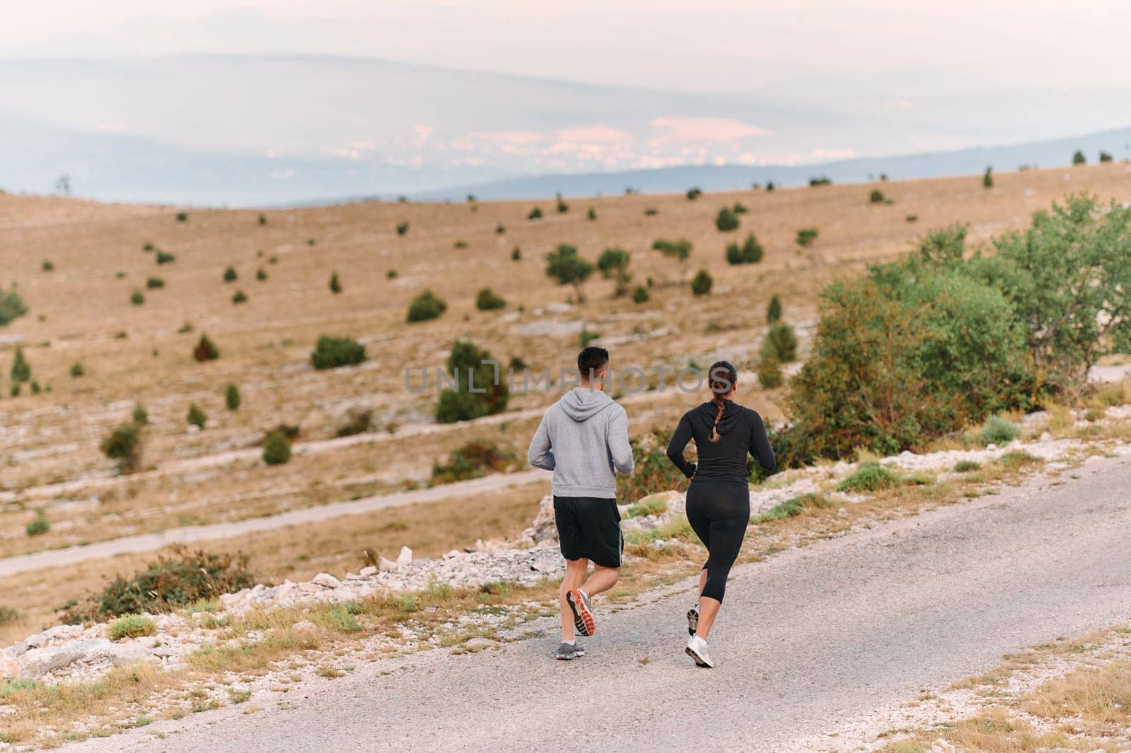 A couple dressed in sportswear runs along a scenic road during an early morning workout, enjoying the fresh air and maintaining a healthy lifestyle.