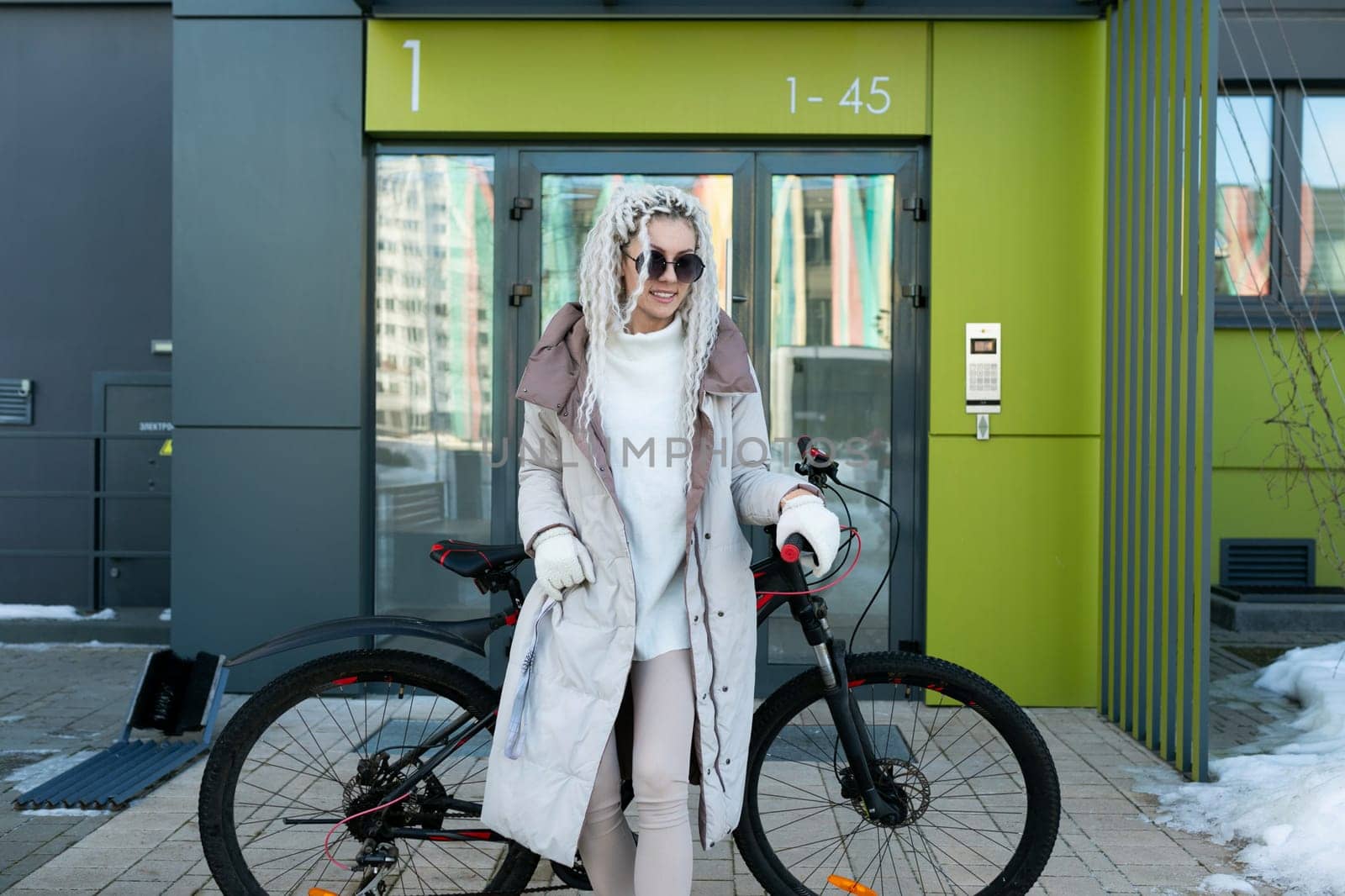A woman is standing next to a bike parked in front of a tall building. She appears to be taking a break or checking her surroundings. The scene captures urban life and transportation in a city setting.