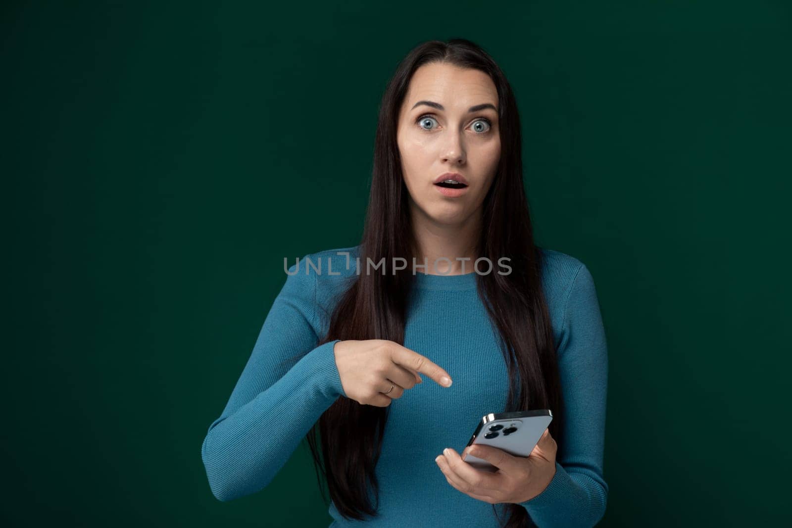 A woman is pointing at a cell phone with a surprised look on her face. She appears to be reacting to what she sees on the screen, showing astonishment or disbelief.