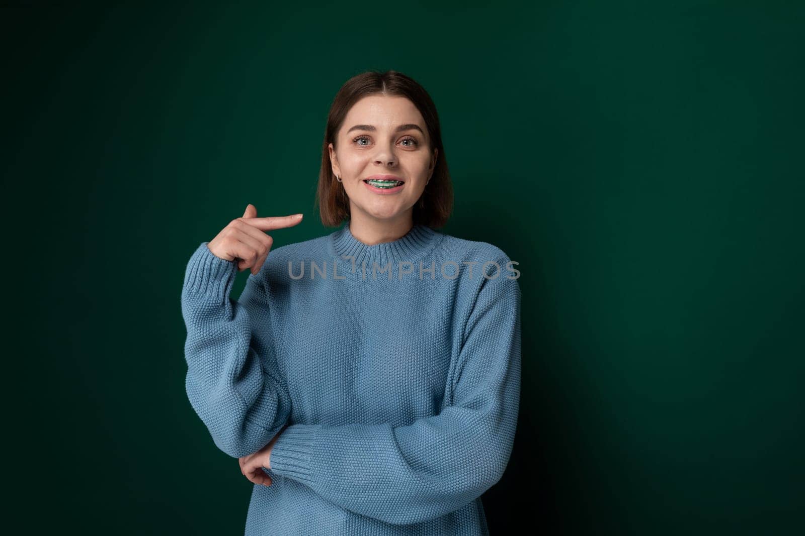 A woman wearing a blue sweater is pointing at an unseen object with a serious expression, indicating curiosity or interest. She is standing against a neutral background, focusing all attention on her gesture.