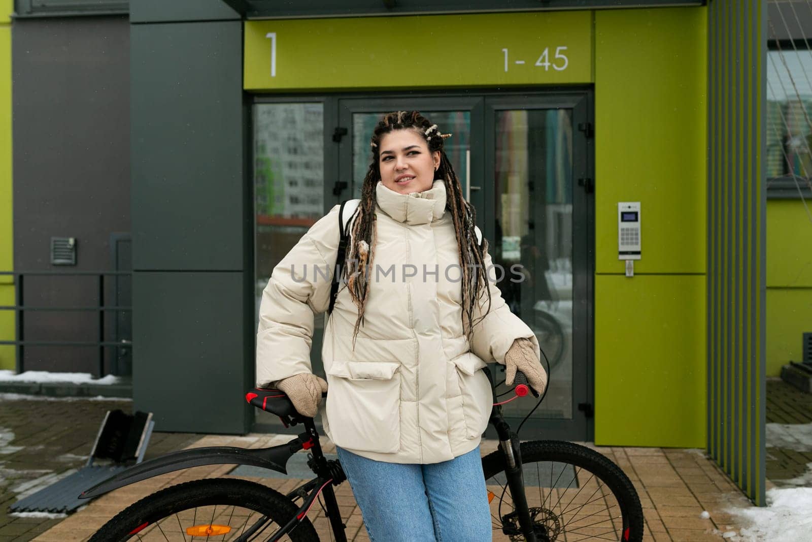 A woman is standing next to a bike in front of a building. She appears to be taking a break or preparing to ride her bike. The building behind her is nondescript, serving as a backdrop to the scene.