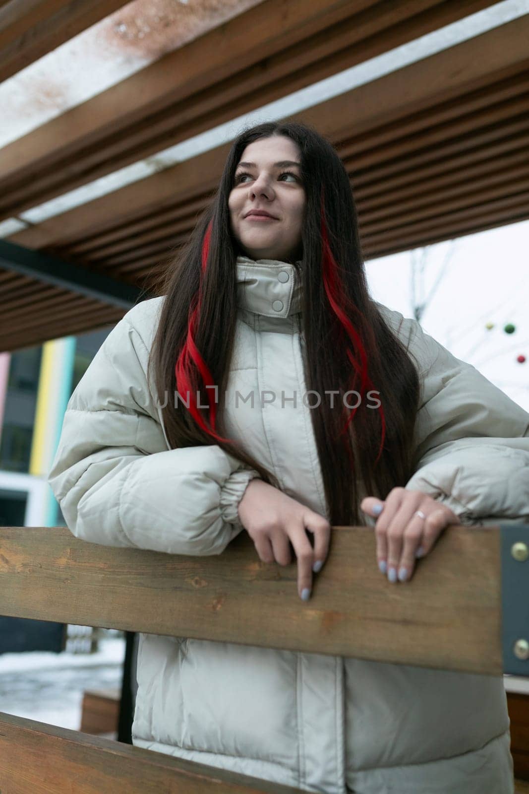 A woman with long hair is leaning on a wooden bench in a park. She appears relaxed and thoughtful, enjoying a moment of rest. Her flowing hair contrasts with the solid structure of the bench.