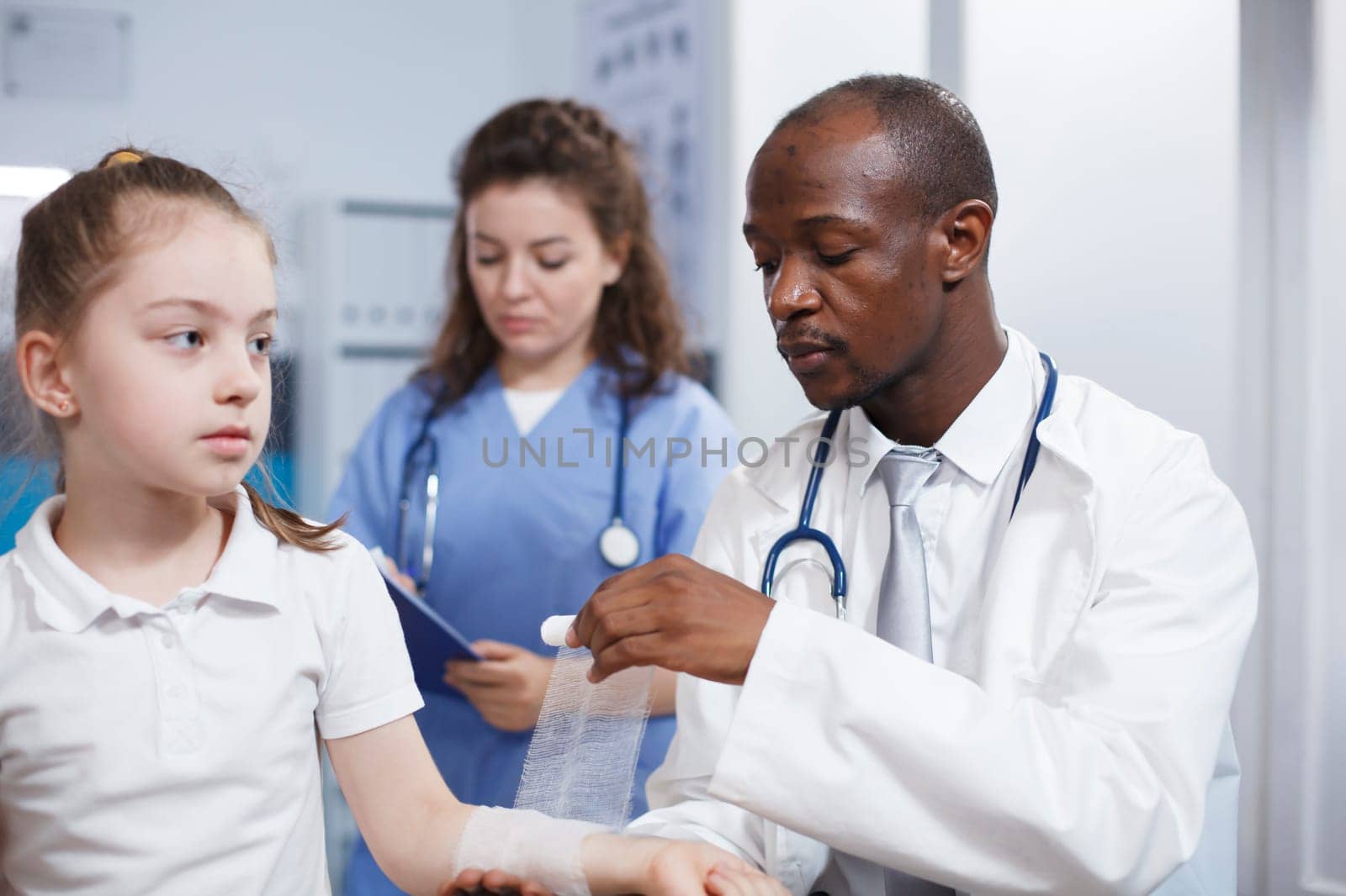 Close-up of a patient arm injury being methodically wrapped in sterile bandage by multicultural medical workers. Young child is being treated by a doctor and a nurse in this photograph.