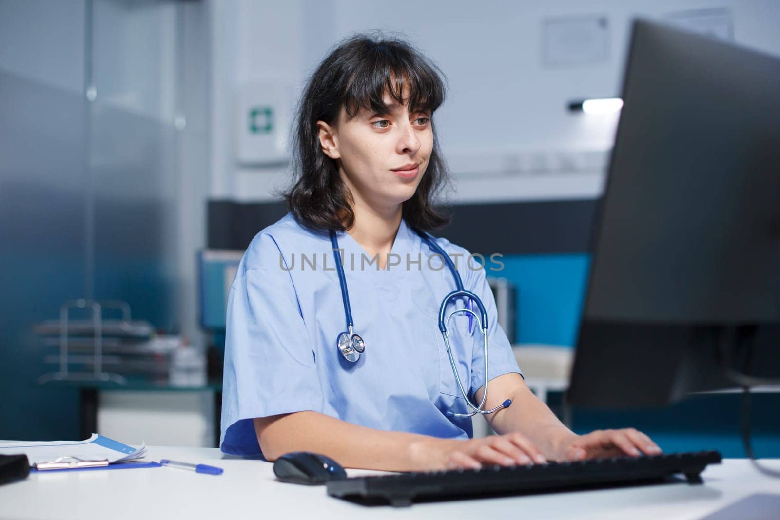 Female practitioner in a modern clinic office using a keyboard to type medical notes on a computer. Caucasian nurse with a stethoscope updating patient information on desktop pc.