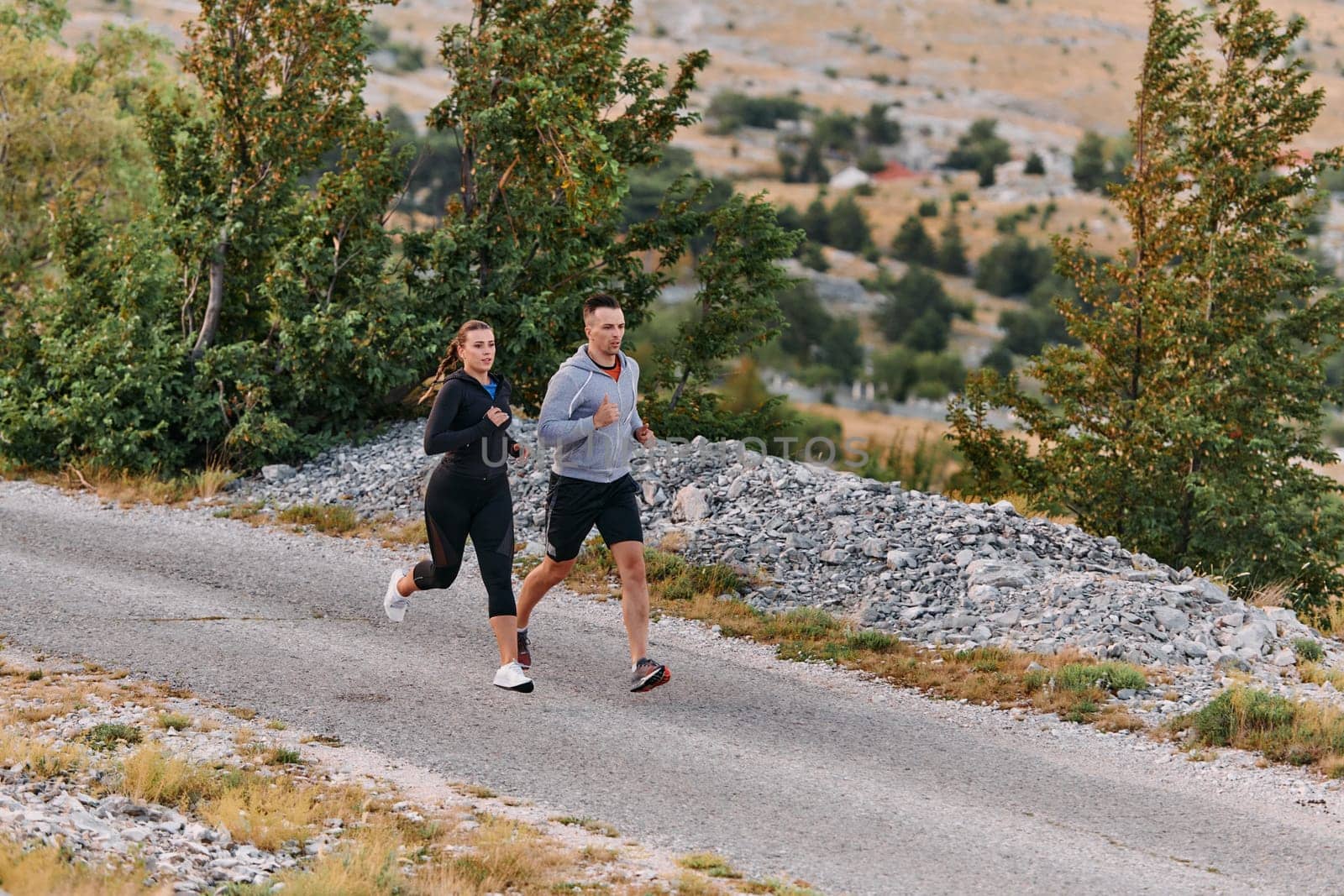 A couple dressed in sportswear runs along a scenic road during an early morning workout, enjoying the fresh air and maintaining a healthy lifestyle.