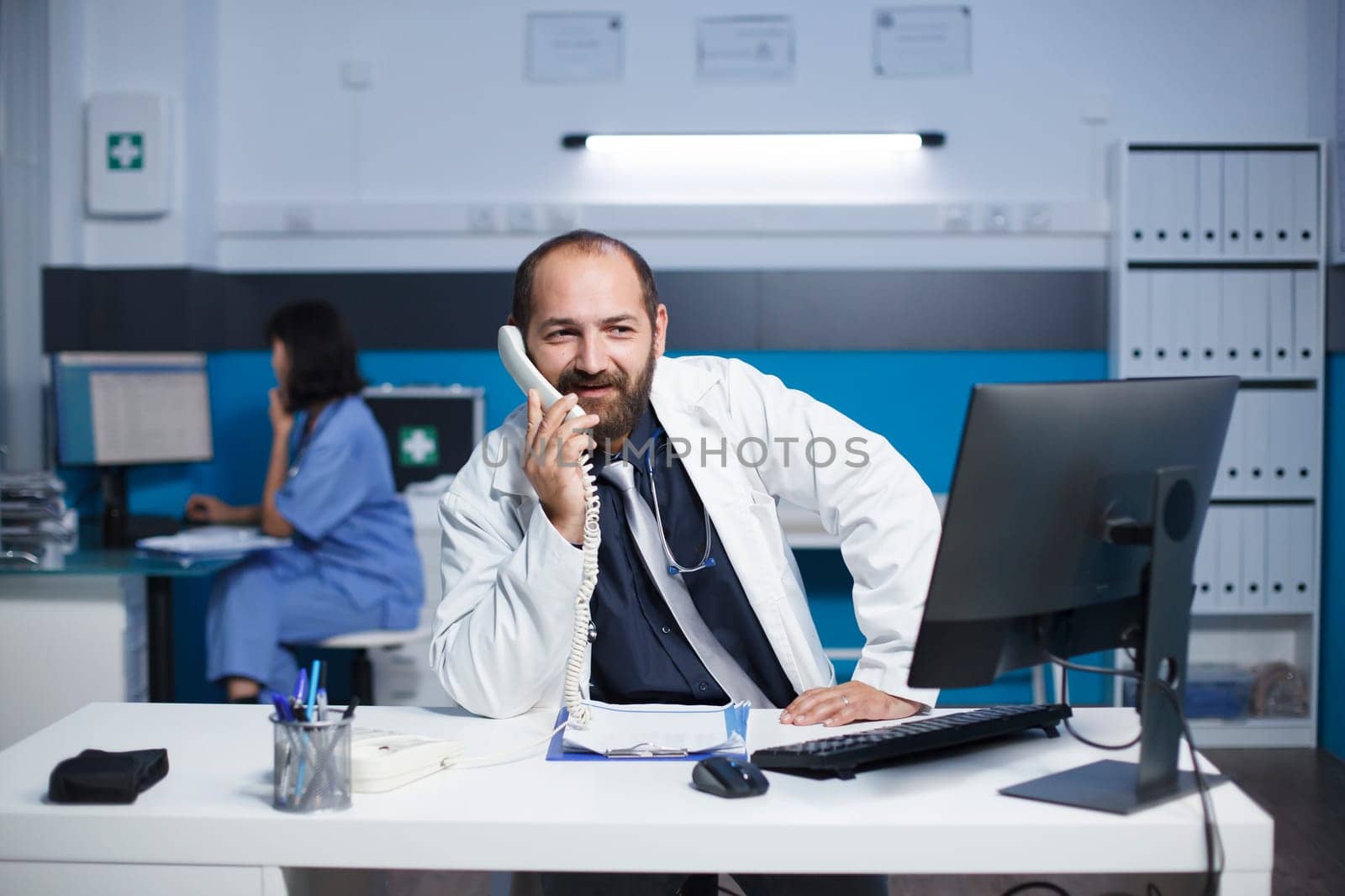 Caucasian male doctor seated at the office desk talking on the telephone with other healthcare specialists. Man wearing a lab coat is using a landline phone to speak with a hospital receptionist.