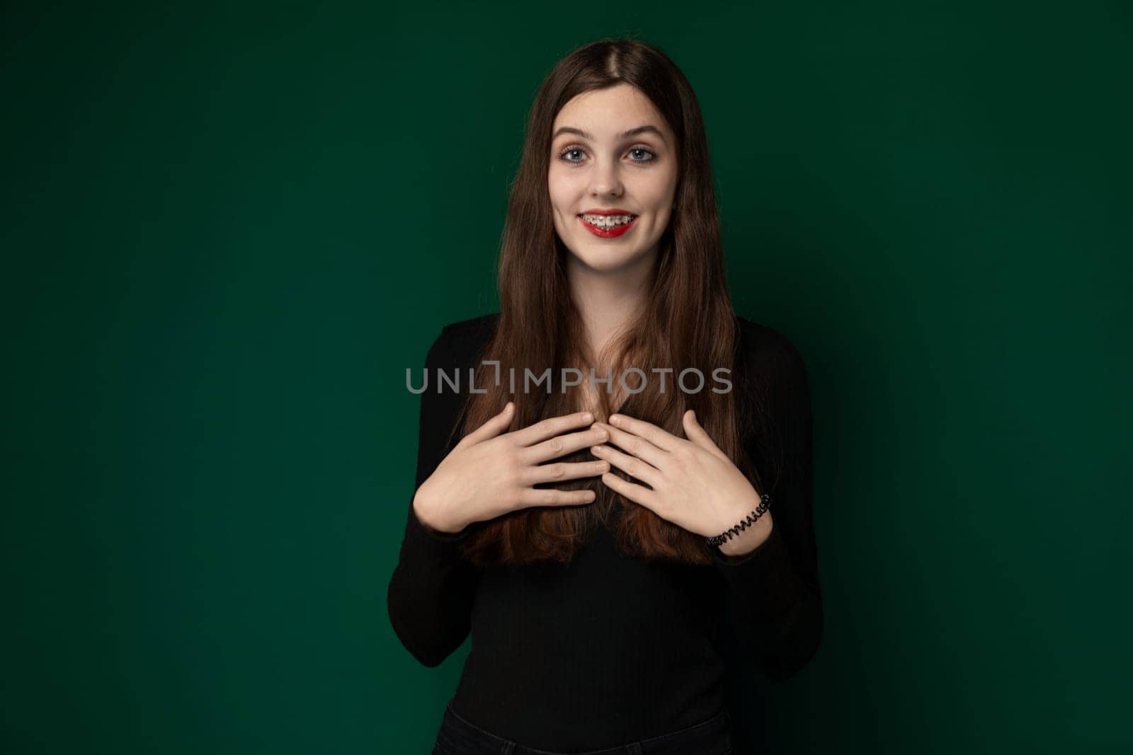 A woman is standing upright in front of a solid green wall. She appears to be looking straight ahead with her hands relaxed by her sides. The simplicity of the scene draws attention to the contrast between the womans figure and the vibrant green background.