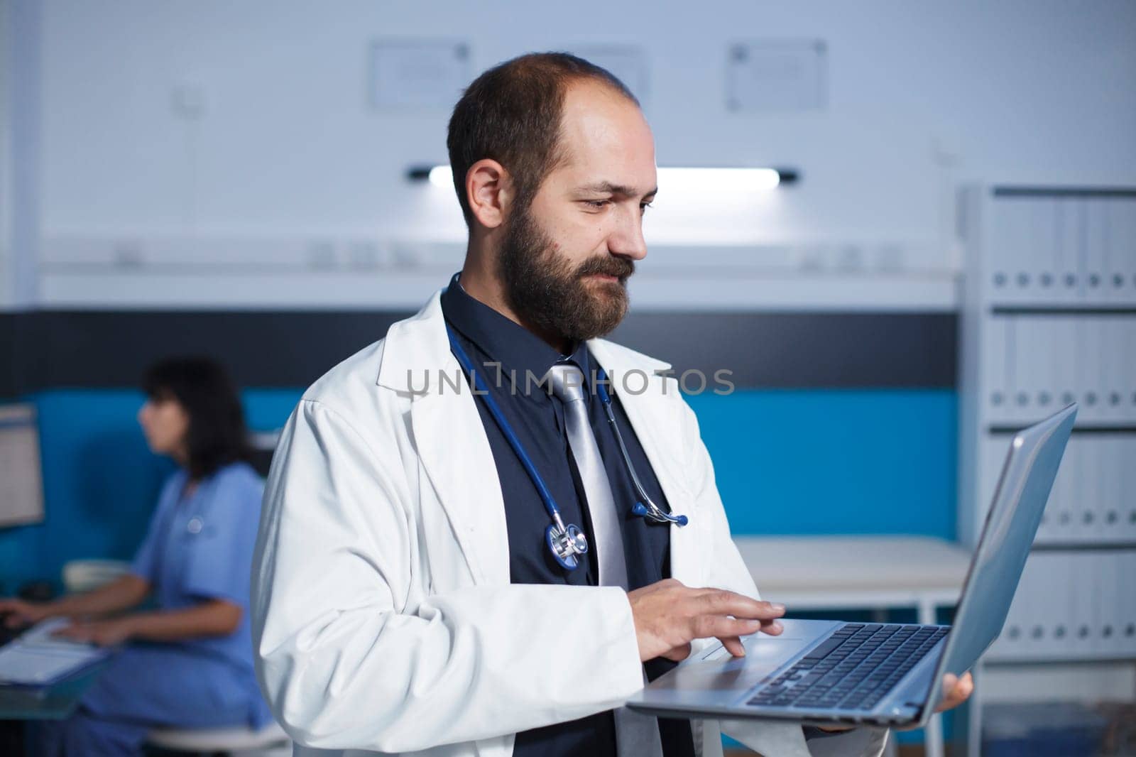 Caucasian doctor is standing and searching for something on his personal laptop, in medical office. In background, nurse using desktop pc with detailed focus on male physician holding a minicomputer.