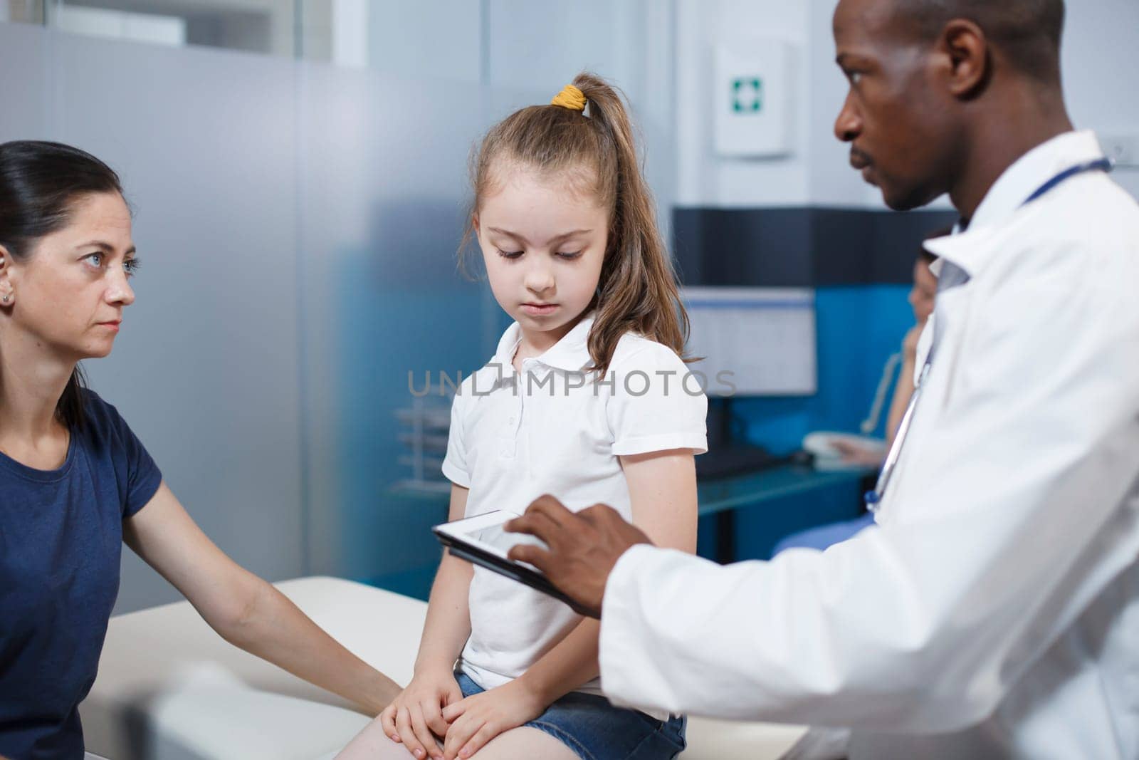 Black man wearing lab coat consults with girl patient and mother in clinic office. African American physician using digital tablet while providing medical advice to caucasian woman and her daughter.