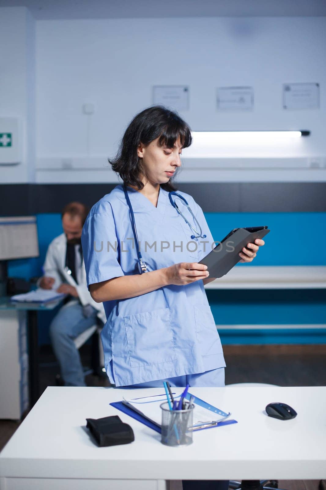 Female medical worker looking at tablet in an office while standing next to desk having pens, a clipboard, and a mouse. In the background sits a male physician working on healthcare treatments.