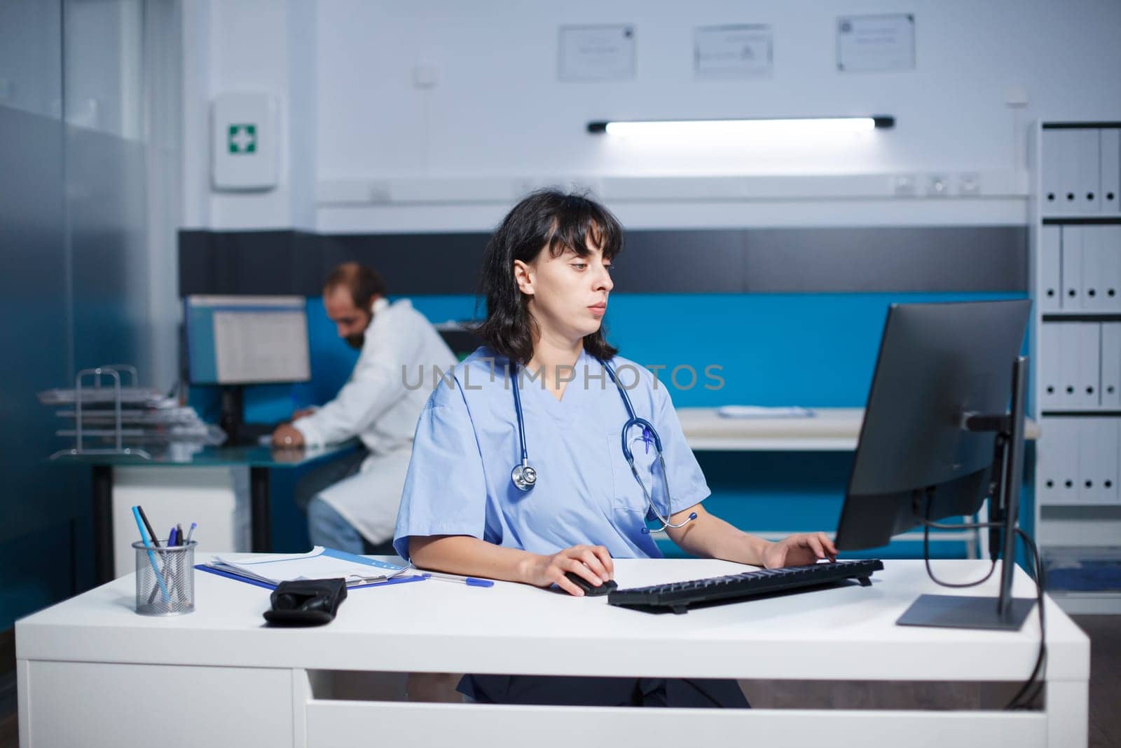 Cucasian practitioner is typing a medical report at a desk in a hospital office late at night. Female nurse preparing medical care while assessing patient illness symptoms.