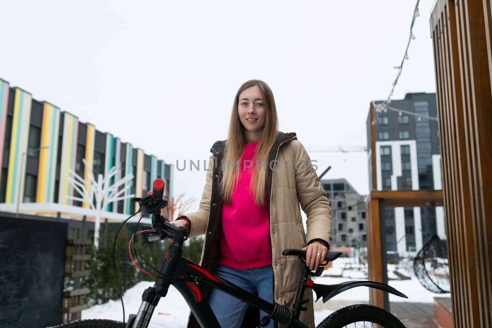 A woman with dark hair is standing beside a bicycle parked in the snow-covered ground. She is wearing a heavy winter coat and boots, looking towards the distance.