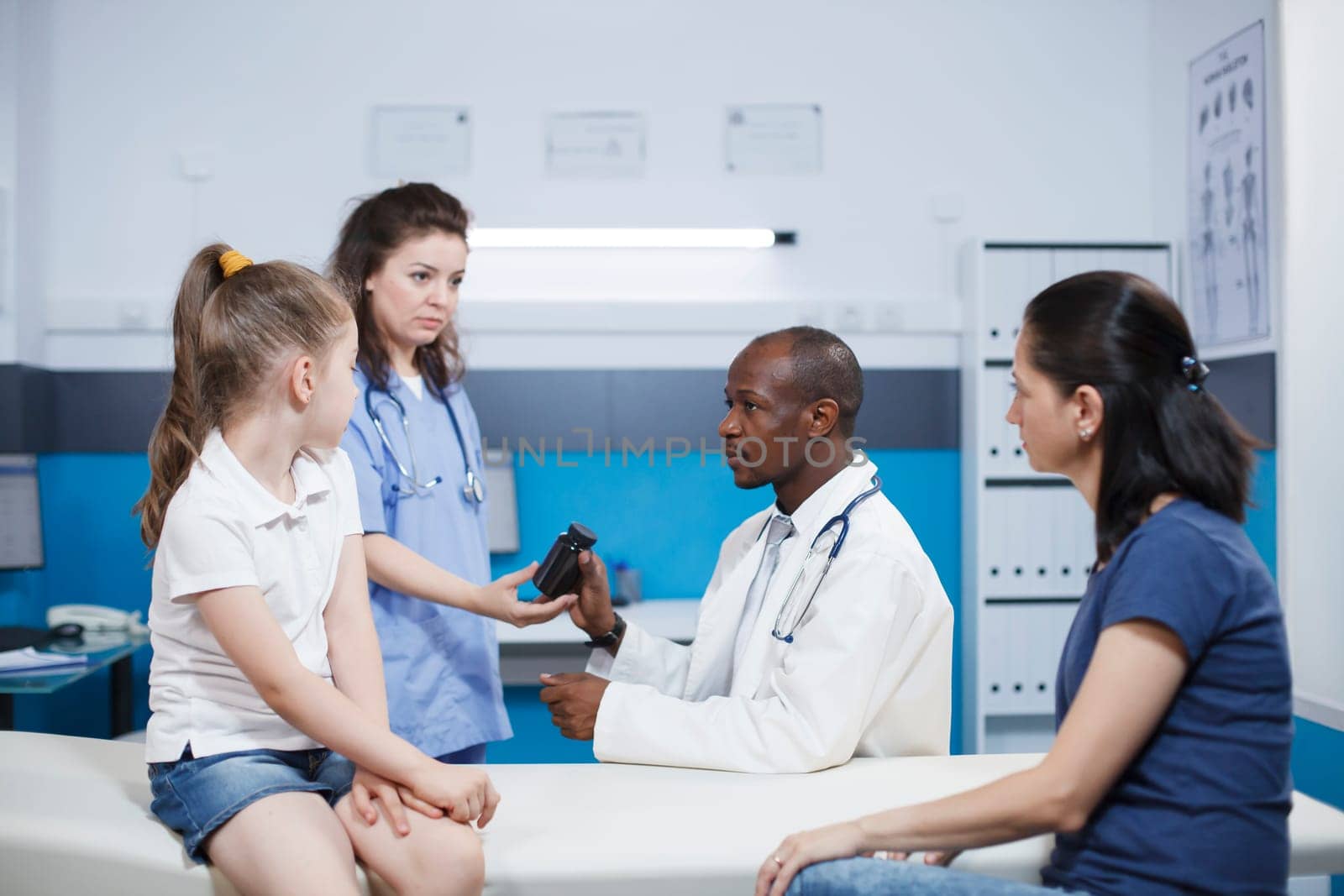 Caucasian mother and daughter in an office consulting with African American man in lab coat and a female nurse. Medical assistant in blue scrubs giving a bottle of medicine to doctor for patients.