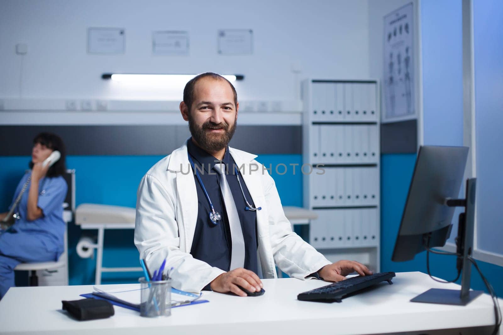 Middle aged doctor smiling at his desk in a medical office. Caucasian male physician appears to be wearing a white lab coat, female nurse is in the background on a call. Portrait shot.