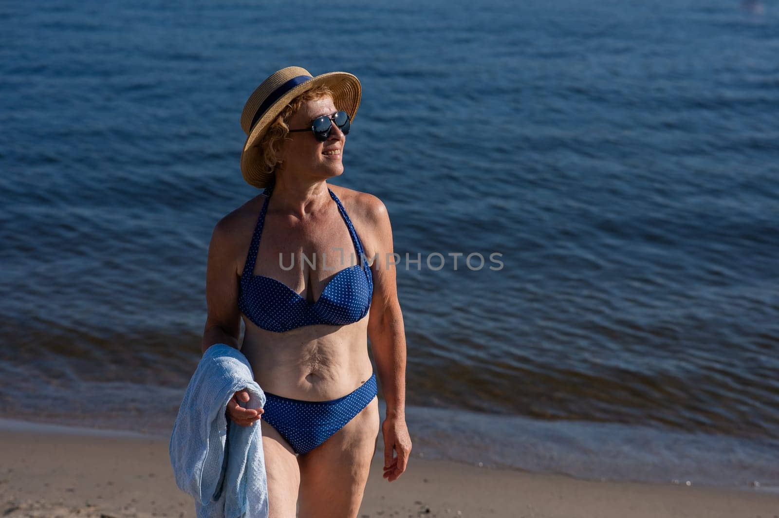 An old woman in a straw hat, sunglasses and a swimsuit is resting on the beach. by mrwed54