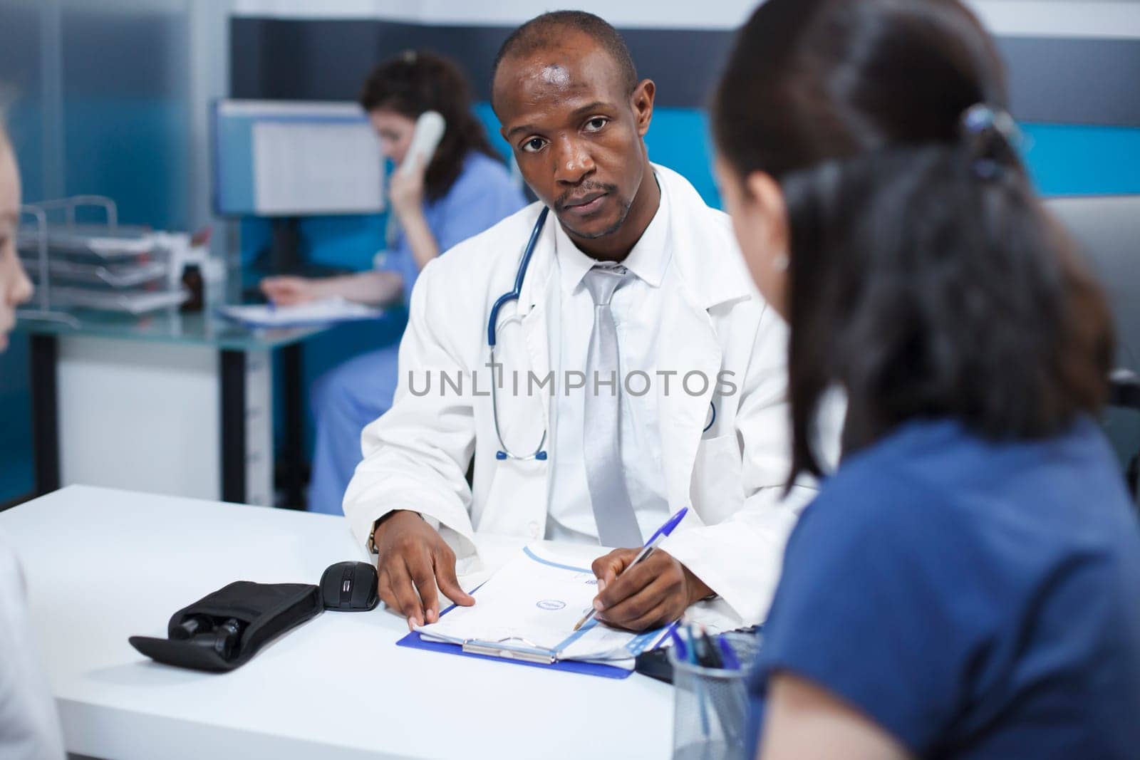 Black man wearing a lab coat, taking notes during a consultation with a female patient at the clinic. African American doctor explains the treatment plan, ensuring proper care.