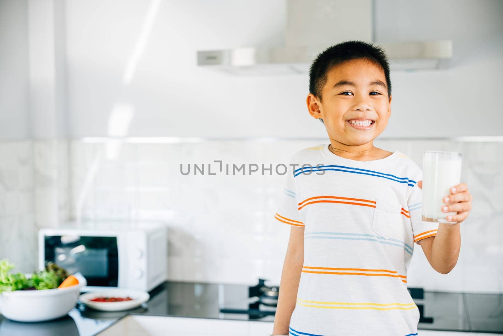 Smiling Asian little boy in kitchen holds milk cup. Portrait of cute son enjoying drink. Joyful child sips calcium-rich liquid, radiating happiness at home, Daily life health care Medicine food by Sorapop