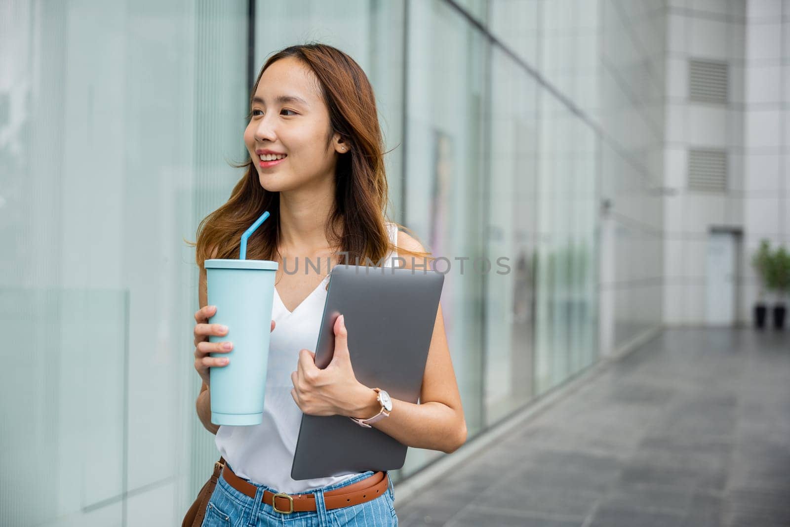 A female entrepreneur with a laptop and tumbler mug water at a shopping mall, using technology to stay connected and productive while on the go.