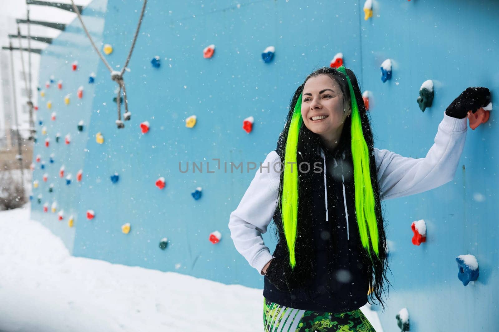 A woman with vivid green hair stands confidently in front of a challenging climbing wall, prepared to tackle the ascent. The colorful hair adds a striking contrast to the rugged terrain behind her.